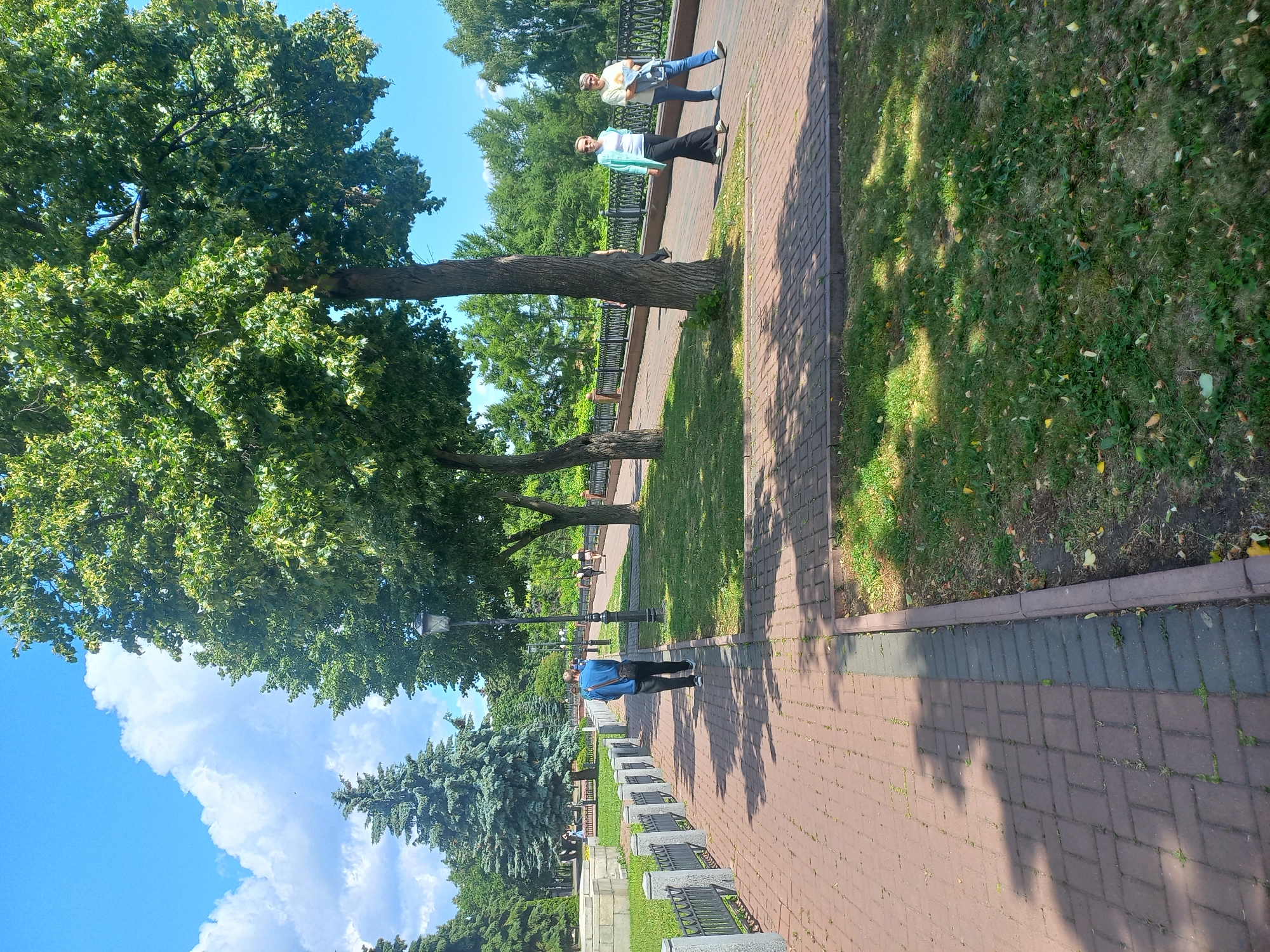 This is a vertical, eye-level, outdoor photograph of a park pathway in Ulyanovsk, Russia. The pathway is paved with red and gray bricks, and lined with a black metal fence and stone pillars. Lush green grass and trees border the pathway. Several people are walking on the path. In the foreground, a man in a blue shirt and black pants walks away from the camera. Further down the path, two women walk towards the camera, one in a teal cardigan and white top, the other in a yellow t-shirt and jeans. The sky is blue with fluffy white clouds. The scene appears to be a sunny day, with shadows from the trees cast across the pathway. The photograph captures a serene atmosphere of a public park.