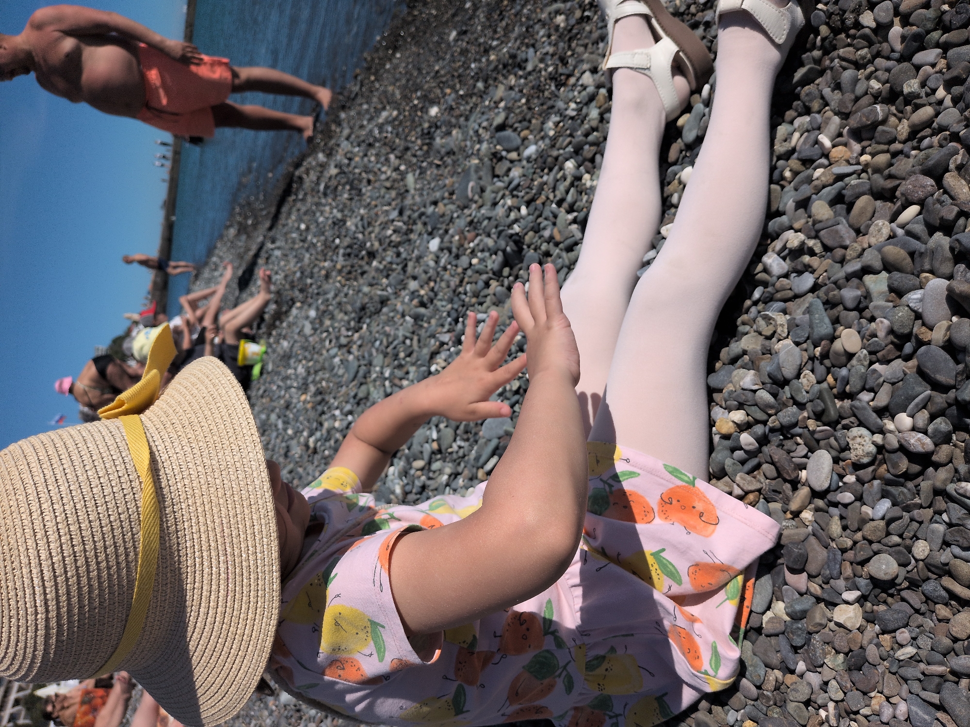 A sunlit, high-angle shot captures a young child lying on their back on a pebbly beach in Sochi, Russia. The child is wearing a straw hat with a yellow ribbon, a pink short-sleeved dress patterned with cartoon lemons and oranges, white tights, and white closed-toe sandals with ankle straps. Their arms are bent with hands slightly raised, suggesting play or relaxation.
The beach consists of numerous grey, brown, and white pebbles of varying sizes. In the background, the calm, blue sea is visible, with several other people enjoying the water and the shore. One adult male, wearing orange swim shorts, stands near the water's edge on the left side of the frame. Other blurry figures are scattered further along the beach.
The bright sunlight indicates it is daytime, likely mid-day, with clear blue skies reflected in the water. The scene evokes a leisurely summer day at the beach. There is no visible text in the image.