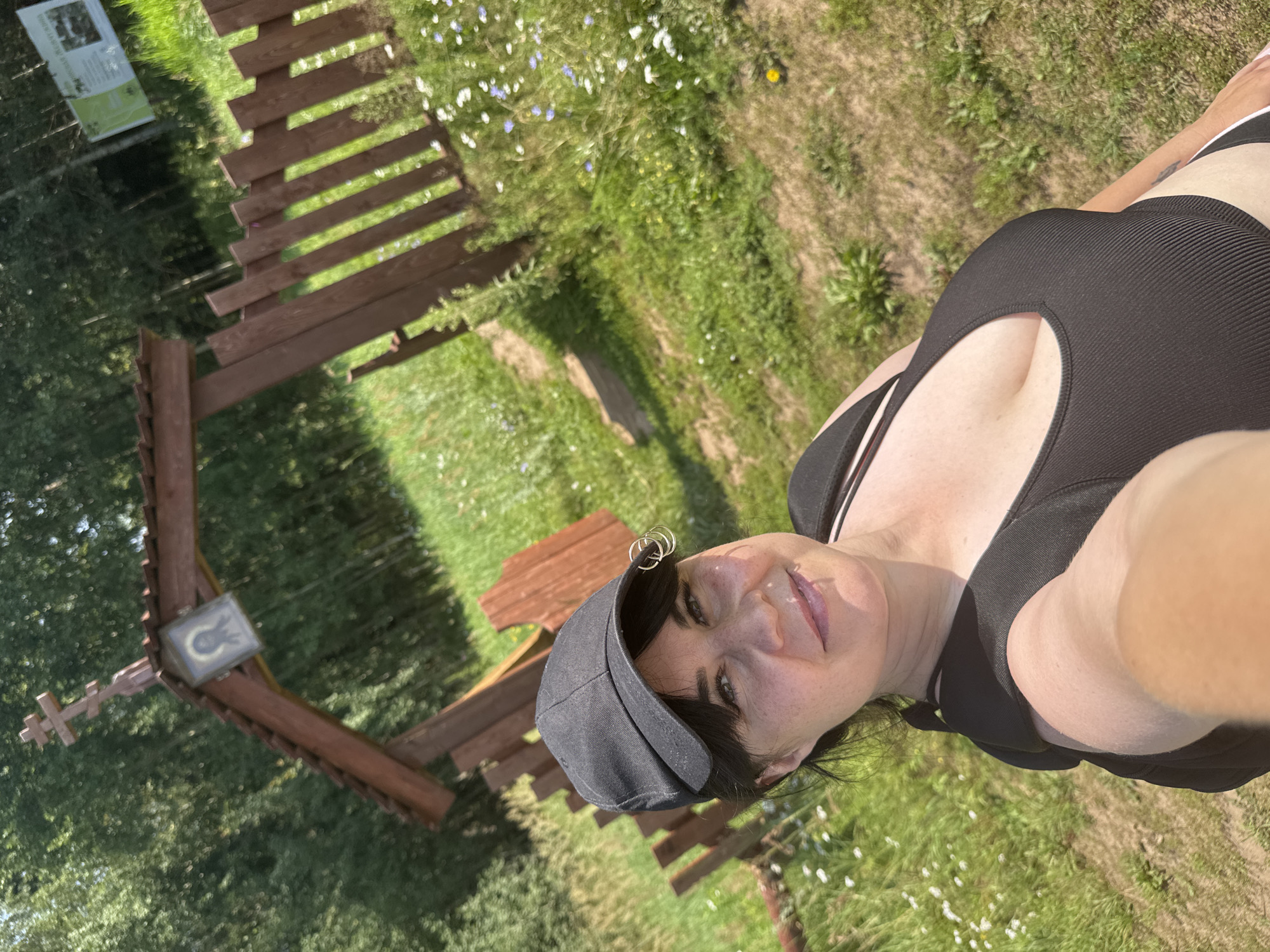 A woman in a black baseball cap and a black ribbed tank top is taking a selfie outdoors. She is wearing large hoop earrings. Behind her, a wooden archway with a cross and an icon of a religious figure stands in front of a wooden fence. The scene is set in a grassy area with scattered wildflowers and trees in the background. A sign with text and images is visible on a pole in the distance. The overall setting appears to be a park or natural area.