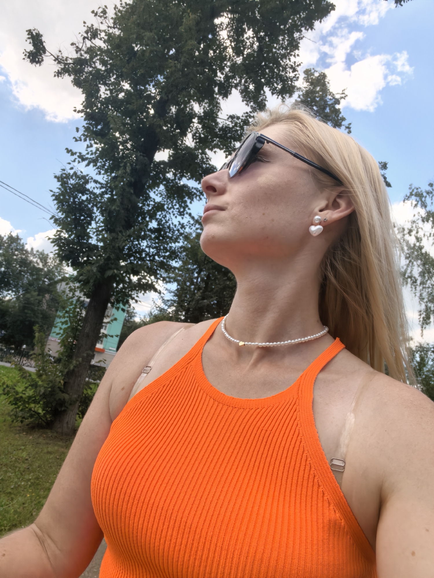 A woman with blonde hair and sunglasses is looking up and to the side. She is wearing an orange ribbed tank top, a pearl necklace with a small gold heart charm, and pearl earrings shaped like hearts. The background shows trees and a blue sky with clouds, suggesting an outdoor setting. The location is identified as Sterlitamak, Russia.