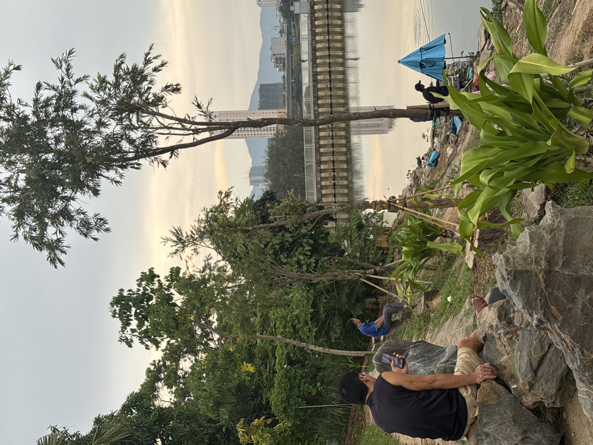 The image depicts a park scene in Đà Nẵng, Vietnam, likely near the Hàn River. In the foreground, a man wearing a black tank top and khaki shorts is sitting on large rocks and looking at his mobile phone. Behind him, another man in a blue shirt is reclining on a bench-like structure. The middle ground features greenery, including trees and shrubs. In the background, the Hàn River reflects the city skyline with its buildings and a bridge. Further along the riverbank, a blue umbrella is visible, indicating a possible fishing spot. The overall scene suggests a tranquil urban park setting.