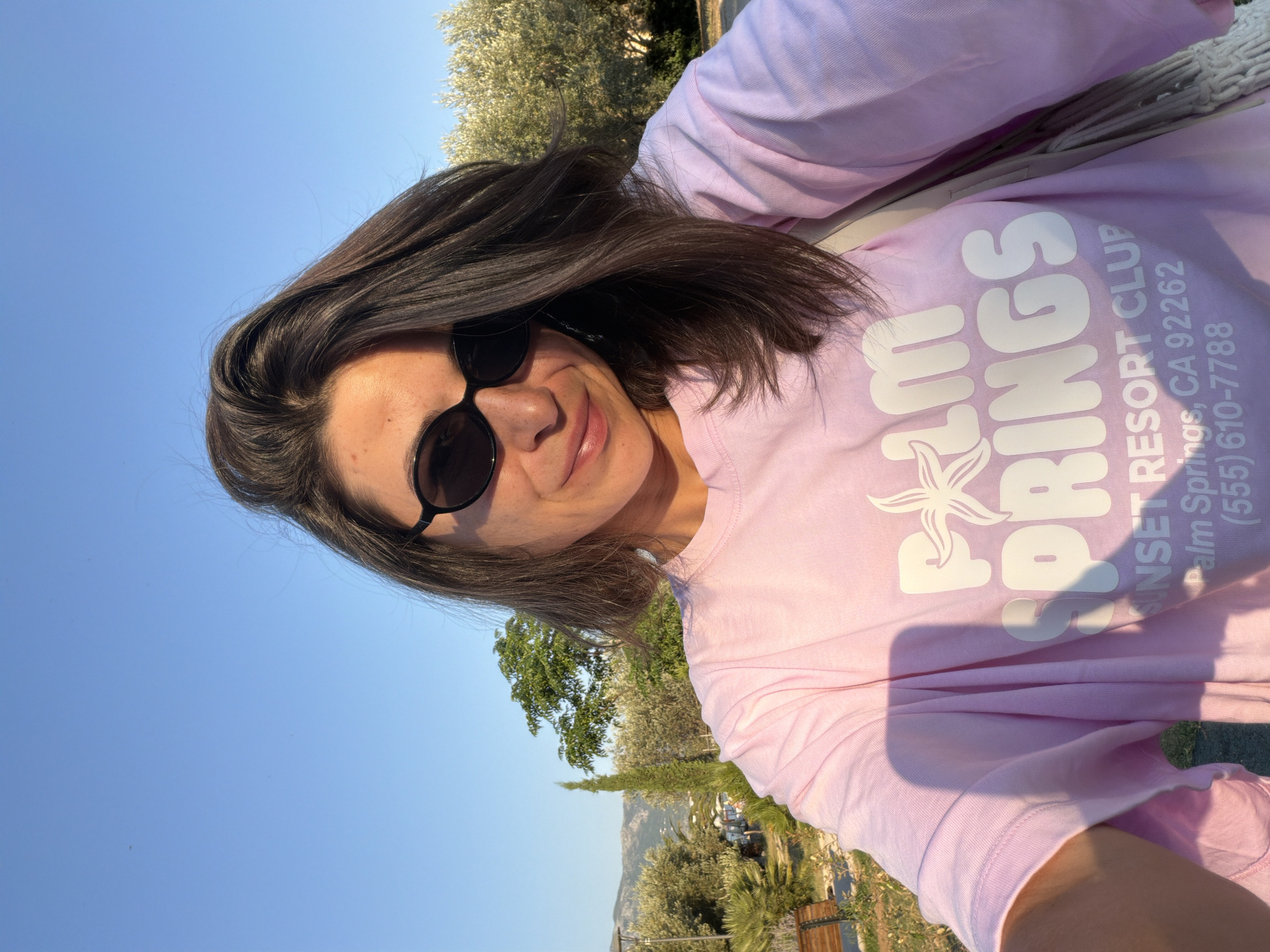 A woman with dark hair and sunglasses takes a selfie outdoors. She is wearing a light pink t-shirt with the words "Palm Springs Resort Club" and a phone number printed on it. Behind her, there are trees, hills, and a clear blue sky. The lighting suggests it is late afternoon. The location is likely near the coast of Budva, Montenegro, given the presence of Mediterranean vegetation and the characteristic hills.