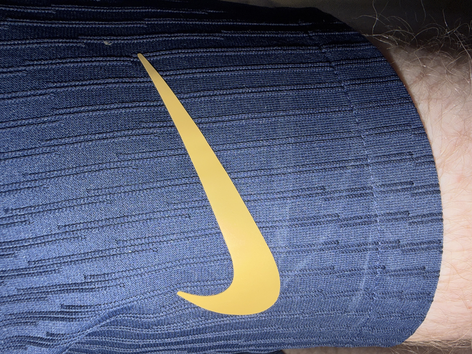The image shows a close-up of a navy blue garment with a ribbed texture and a yellow Nike swoosh logo. The garment is dark blue with vertical lines created by the ribbed fabric. The Nike logo is a bright yellow curved shape, positioned on the left side of the garment, pointing towards the right. The fabric appears to be made of a synthetic material, possibly polyester or nylon. The bottom portion of the image reveals skin with light hair, suggesting the garment is being worn on a person's leg. There are no discernible environmental details as the focus is solely on the garment and the logo. The location is not specified, and the image is a still photograph.