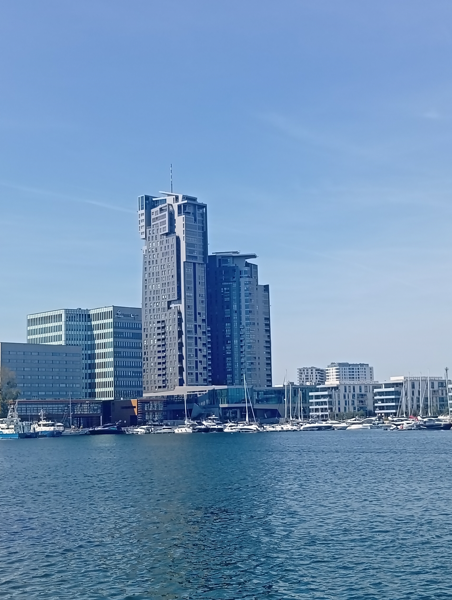The image depicts the waterfront of Gdynia, Poland, with a focus on the city's modern skyline. In the foreground, a body of blue water, likely the Baltic Sea, is visible, with small ripples on its surface. Several boats and yachts are docked along the waterfront, their white sails and hulls contrasting with the blue of the water and sky. Behind the boats, a row of contemporary buildings lines the harbor. These include office buildings with glass facades and several residential towers. The most prominent structure is the Sea Towers, a distinctive pair of skyscrapers, one of which is significantly taller and has a unique stepped design. The sky is a clear, pale blue, with faint wisps of clouds. The overall scene conveys a sense of a well-developed urban harbor and a pleasant, sunny day.