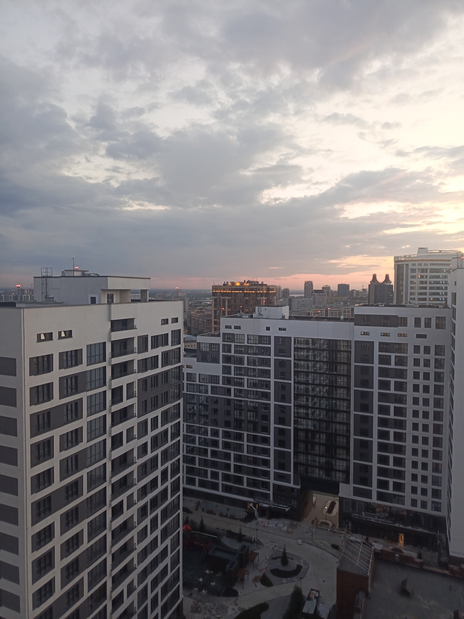 This is a medium-shot, aerial photograph of a city skyline at dusk, likely taken in Novosibirsk, Russia. The foreground features several modern, tall apartment buildings with dark-tinted windows and light-colored facades. These buildings are architecturally similar, suggesting a planned development. The architecture is characterized by repetitive geometric patterns of windows and balconies.
In the mid-ground and background, a dense cityscape is visible, with a variety of buildings of different heights and architectural styles. Some buildings appear to be commercial or office spaces, with illuminated windows. In the distance, the skyline is softened by the haze and the fading light of sunset.
The sky is filled with dramatic clouds, a mix of gray and lighter tones, with hints of pink and orange where the sun is setting. The overall lighting suggests either early morning or late evening. The weather appears to be overcast, contributing to the subdued colors of the scene.
There are no people visible in the image, but there are some indications of ongoing construction or development on the ground level, with what appear to be construction materials and machinery. The scene evokes a sense of urban development and the transition from day to night. No text is discernible in the image.