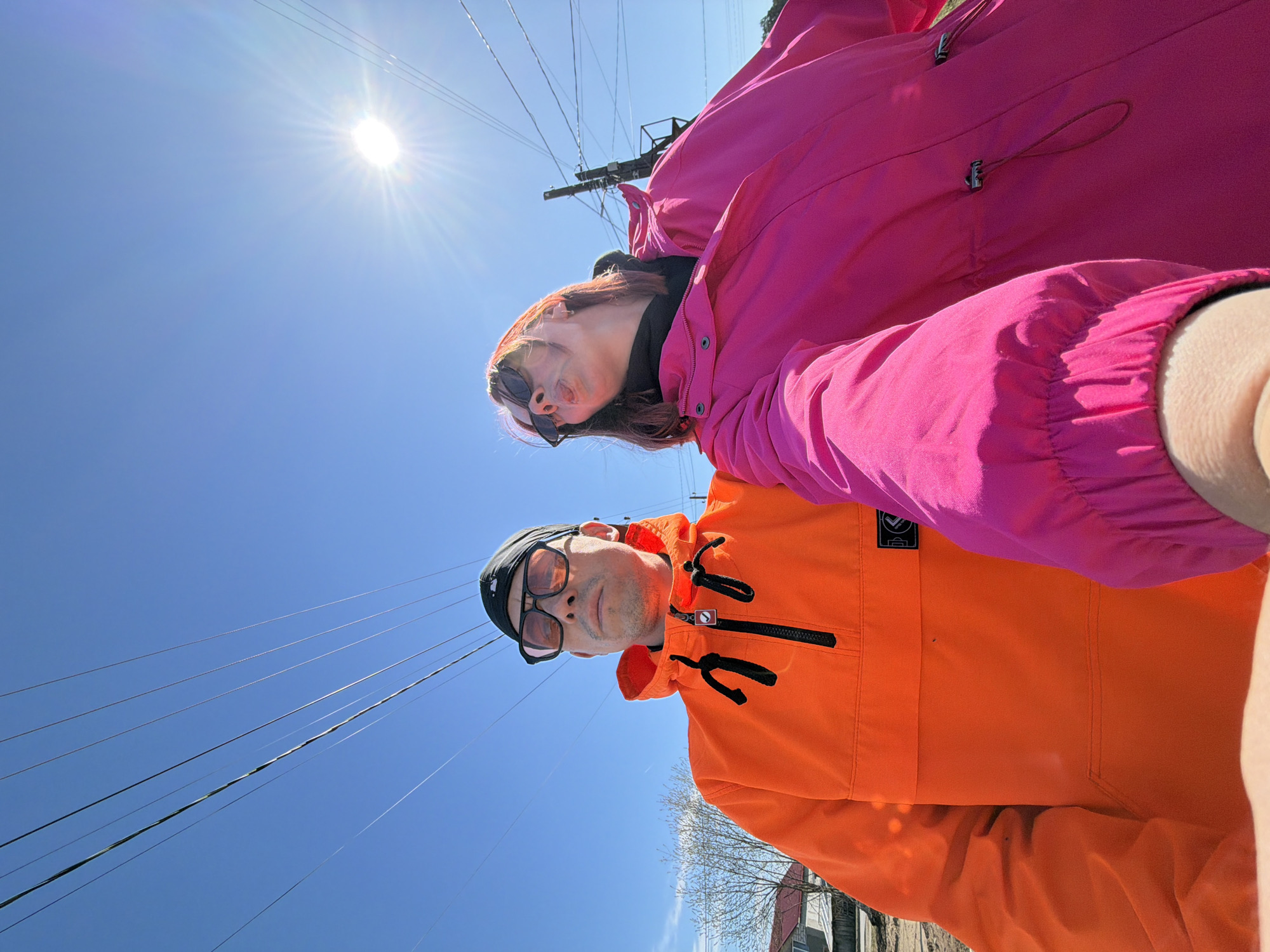 This low-angle selfie, rotated ninety degrees clockwise, captures two individuals looking upwards into a clear blue sky under bright sunshine. The photograph appears to be taken by the woman, whose arm is visible on the right edge.
On the left side of the image (or top if viewed upright), a woman with reddish-brown hair is wearing sunglasses pushed down on her nose, a dark inner layer, and a vibrant hot pink jacket. Her head is tilted slightly back, and she looks towards the camera with a neutral expression.
To her right (or below her if viewed upright), a man is featured wearing a black cap, dark-rimmed sunglasses with tinted lenses, and a bright orange anorak-style jacket with black zippers and drawstrings. He also gazes towards the camera. On his orange jacket, a small black patch with an indistinct white logo is visible on the upper left chest.
The background is dominated by a brilliant clear blue sky, with the sun appearing prominently in the upper left corner, casting strong rays. Multiple black power lines stretch diagonally across the sky, connected to utility poles or structures, one of which is partially visible behind the woman. In the lower right background, bare tree branches suggest a season like early spring or late autumn. Faint outlines of buildings can be seen in the distance at the very bottom right.
The scene depicts a bright, sunny day, likely midday or early afternoon given the sun's intensity and position. The clear sky and light jackets suggest pleasant weather. The setting appears to be an urban or suburban street in Chaikovsky, Russia, characterized by the visible infrastructure of power lines and distant buildings. The overall mood conveyed by the subjects is relaxed.