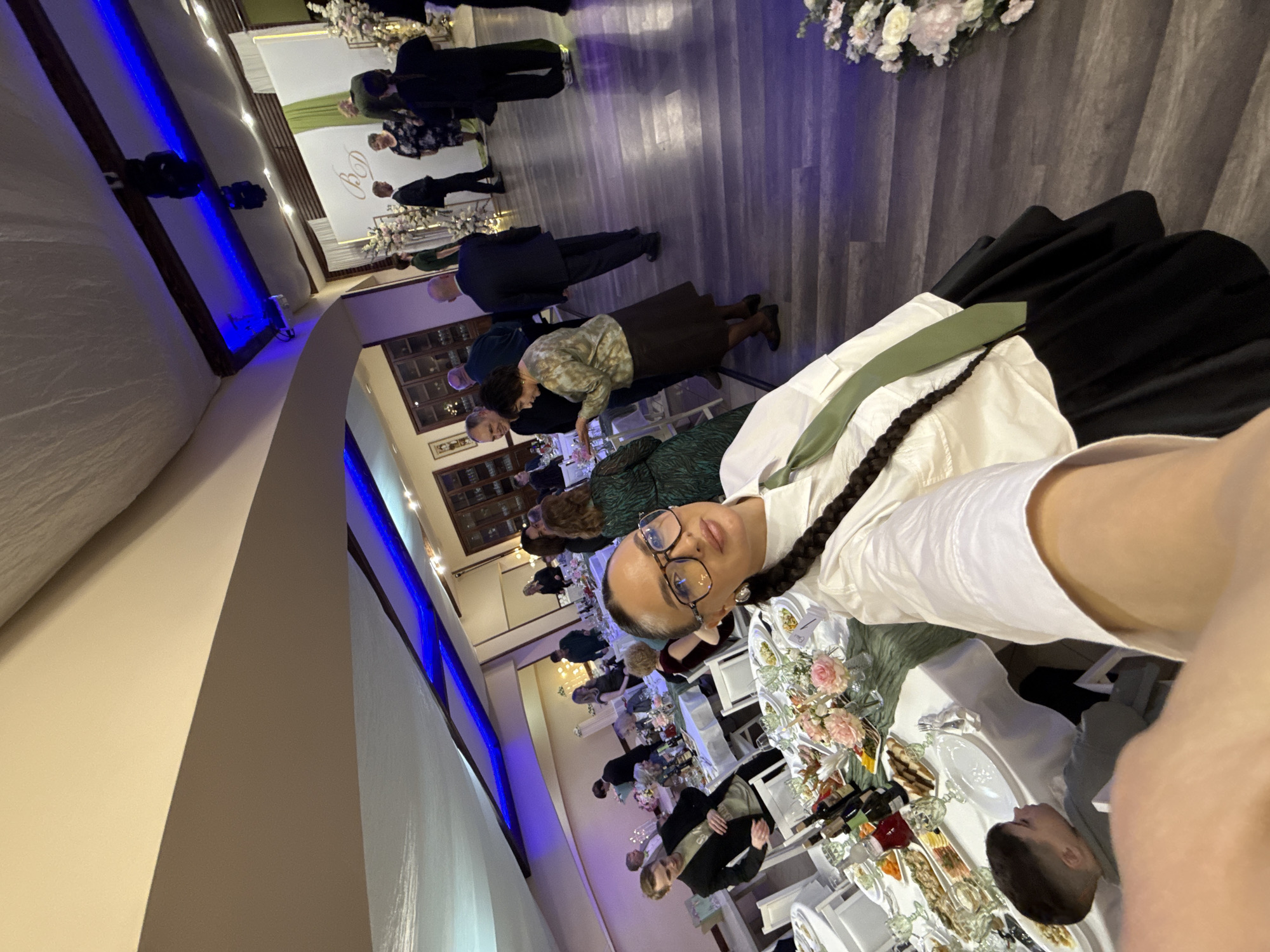 The image captures an indoor celebratory event, likely a reception or banquet, taking place in Chaikovsky, Russia. The scene is dominated by a person taking a selfie in the foreground, looking directly at the camera. This individual has dark hair styled in a long braid, wears glasses, a white collared shirt, and a green tie or sash.
In the immediate foreground, a table is elaborately set with a white tablecloth, white plates, wine glasses, and various dishes of appetizers and snacks. A floral centerpiece with pink and white flowers adds a decorative touch. Part of a person's head, seated at this table, is visible in the lower right corner.
The wider scene reveals numerous other guests, dressed in semi-formal attire such as suits and dresses, mingling and seated at similar white-linened tables throughout the hall. The venue features a wooden floor and light-colored walls, with prominent architectural elements like a large arched opening on the left. A distinctive blue LED strip light runs along the bottom edge of an upper wall section, casting a modern glow.
In the far background, behind the standing guests, a white banner or backdrop is visible, featuring large, intertwined calligraphic initials, possibly "D" and "G" or similar, suggesting a personalized event like a wedding or anniversary. Floral arrangements are also present near this backdrop.
The artificial lighting, including spotlights and the blue LEDs, indicates that the event is taking place in the evening or at night. The overall atmosphere appears social and festive, consistent with a special occasion. There is no visible weather information as it is an indoor setting.