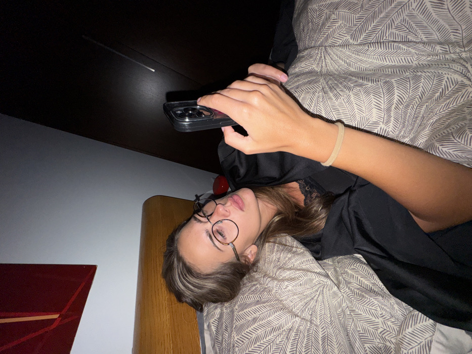 A young woman with round glasses lies in bed, angled toward the viewer. She holds a smartphone above her, presumably looking at the screen. She wears a black silky robe over a lace top. The bed has patterned grey and white sheets. To the left, a red abstract painting hangs above a wooden nightstand. Overhead, a dark ceiling adds to the dimly lit atmosphere.