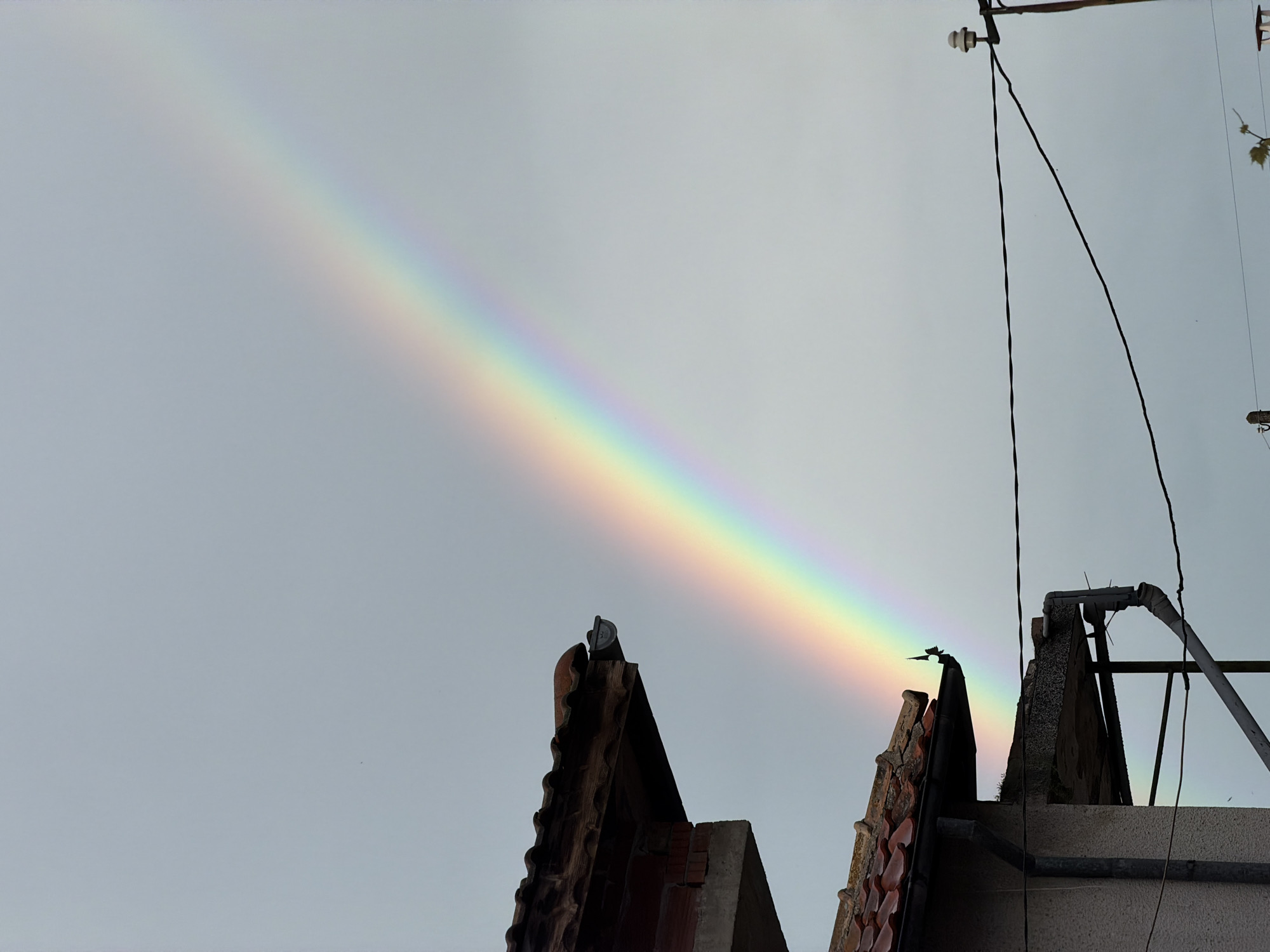 A rainbow is partially visible over a building in Burgas, Bulgaria. The rainbow spans from the upper left to the center right of the frame. The building has an angular roof with dark tiles and concrete structures. Electrical wires and metal poles are also visible atop the building. The sky is overcast.