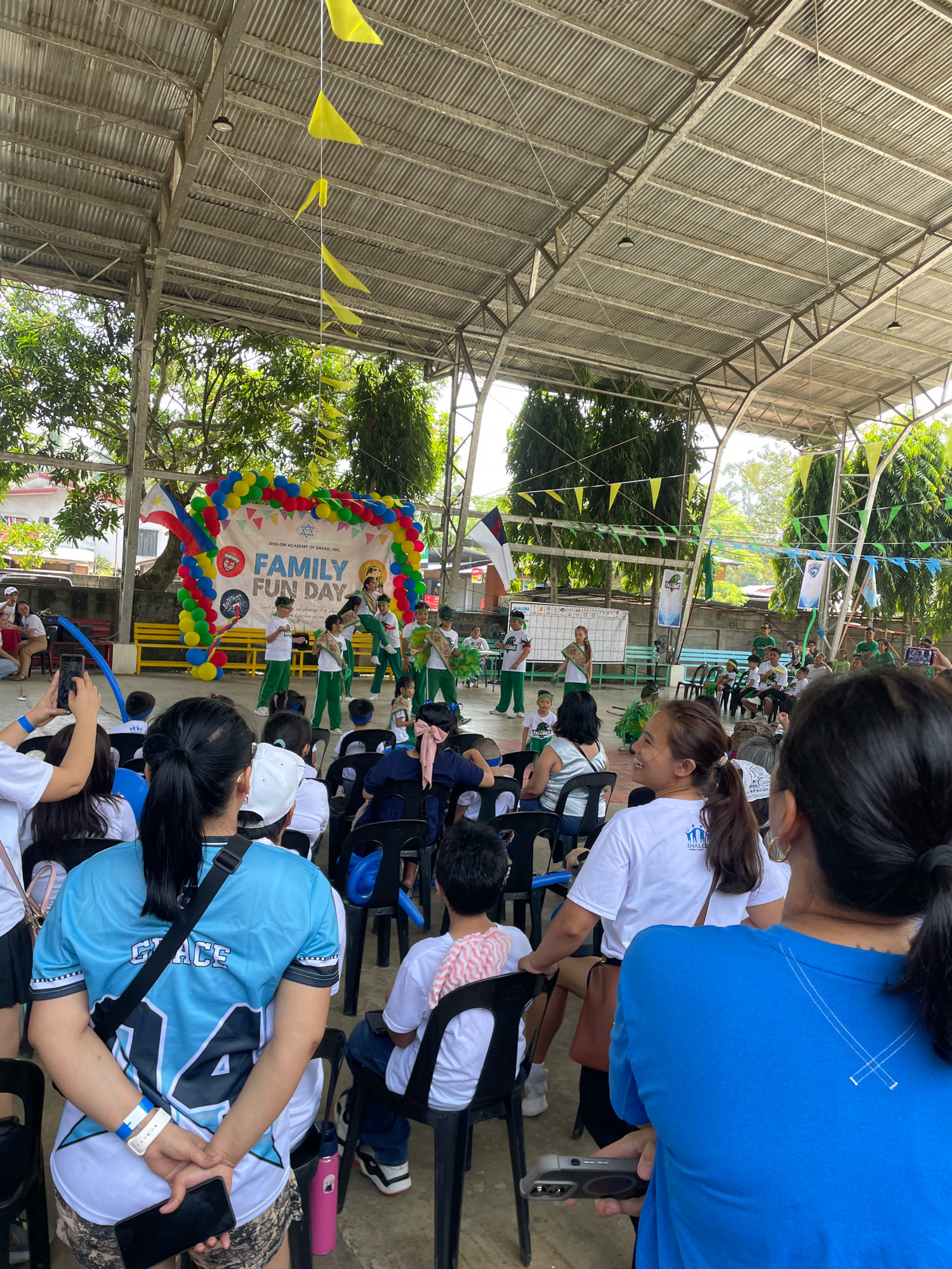 The image depicts an outdoor event at Shalom Academy of Davao City, Philippines, identified by a banner that reads "SHALOM ACADEMY OF DAVAO, INC. FAMILY FUN DAY." The event appears to be a school function held under a covered outdoor court or gymnasium with a corrugated metal roof supported by metal trusses. In the background, a stage is set up with a colorful balloon arch and a banner. Several children, some in green and white uniforms, are performing on stage. Spectators, including adults and children, are seated on black chairs, facing the stage. Some attendees are holding up phones to capture the event. The environment includes trees and buildings visible beyond the covered area, suggesting a semi-urban or school campus setting. The weather appears to be bright and sunny, with light casting shadows. Yellow and blue triangular flags are strung across the ceiling of the covered area, adding to the festive atmosphere.