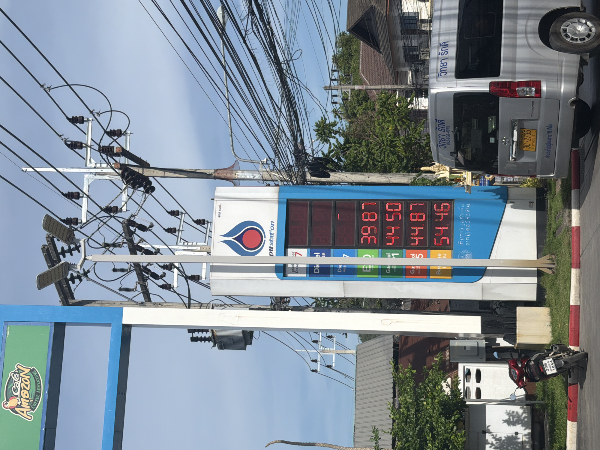 This image depicts a street scene in Baan Mae Nam, Thailand. A PTT gas station sign displays fuel prices, with "Diesel" priced at 39.87, 44.50, and 44.87. Another fuel type is listed at 54.46. A red scooter is parked on the side of the road near the gas station. A silver minivan with Thai license plates is also visible, bearing the text "วิทยา รักษ์" and a phone number. The environment features utility poles with numerous wires and a clear sky. A green sign for "Cafe Amazon COFFEE ADVENTURE" is also present. The time of day appears to be daytime.