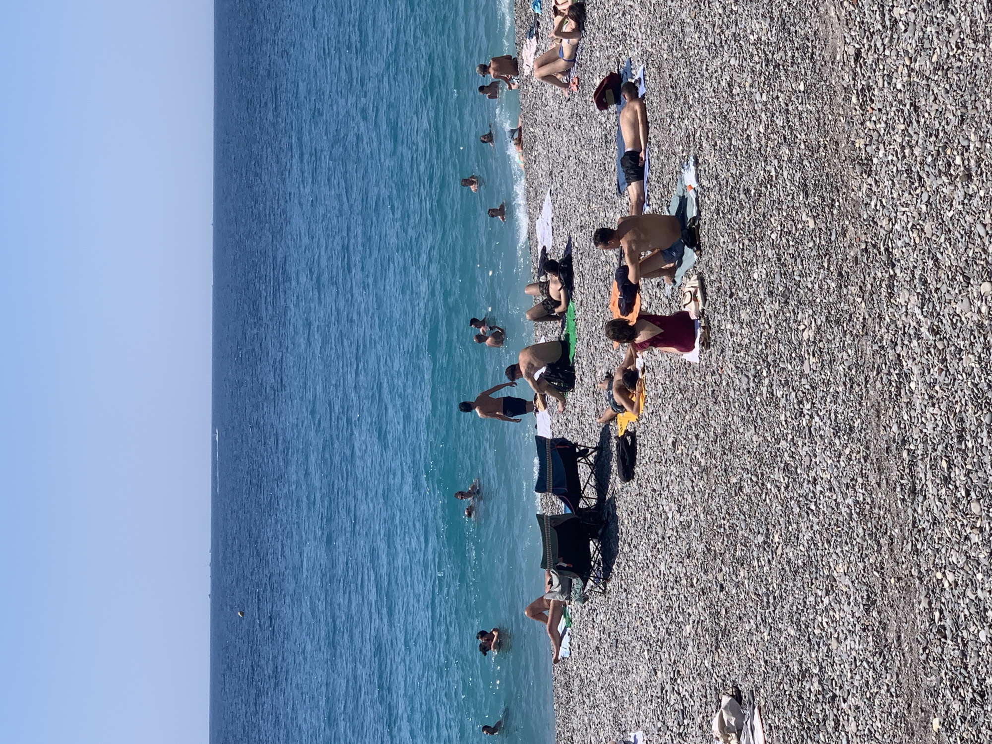 A view of a crowded pebble beach on a sunny day in Nice, France, with the blue Mediterranean Sea stretching to the horizon. Numerous people are sunbathing on towels and chairs on the shore, while others are swimming and wading in the turquoise water. Some boats are visible in the distance on the sea. The scene is bustling with activity as people enjoy the beach.