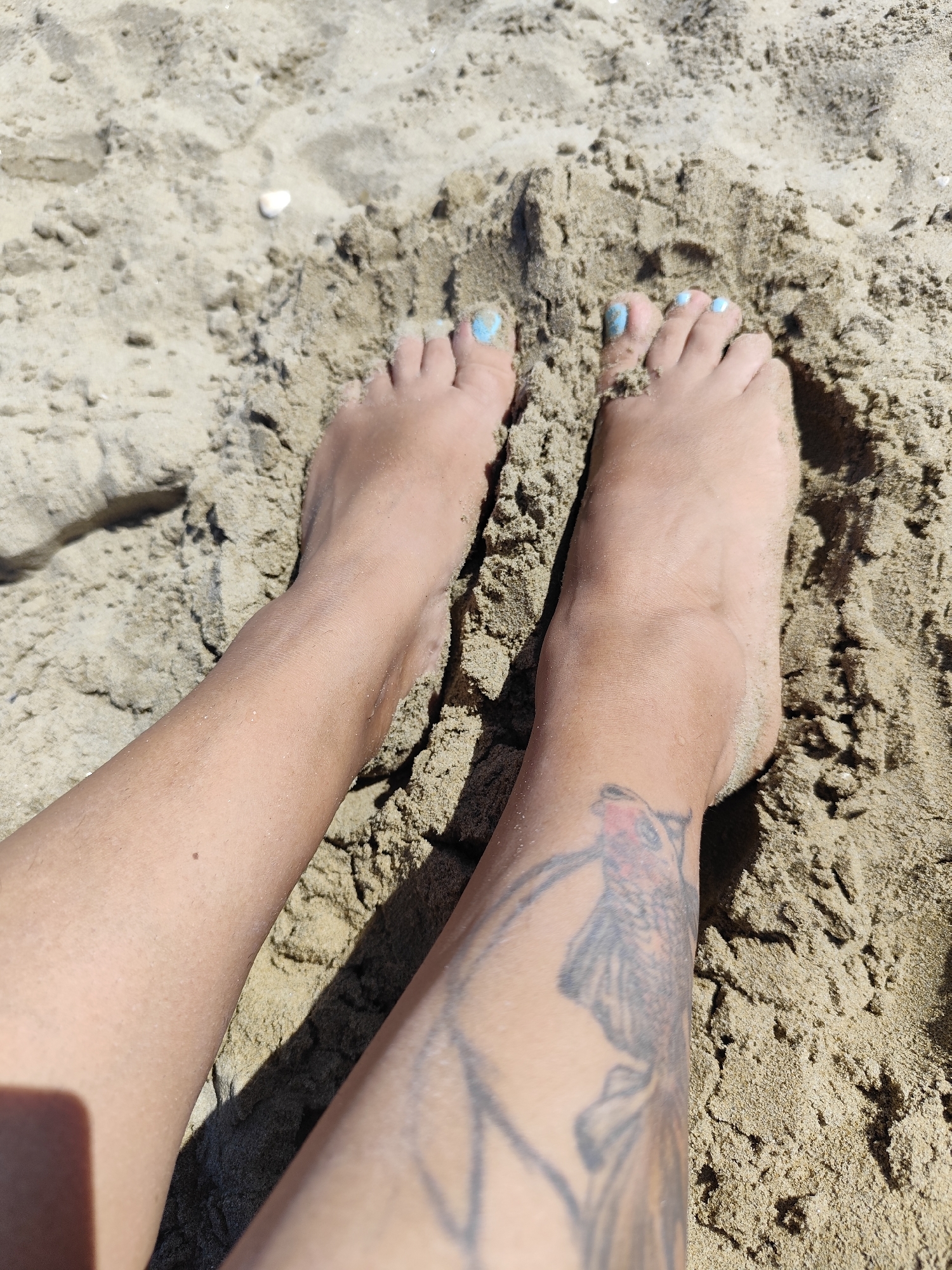 The image shows a close-up view of a person's legs and feet partially buried in sand. The sand appears to be light brown and damp in some areas. The person has light skin and their toenails are painted light blue. There is a tattoo of a bird on the left leg. The scene is outdoors, consistent with a beach setting. No specific landmarks or structures that would indicate the city of Rimini, Italy are visible in the image.