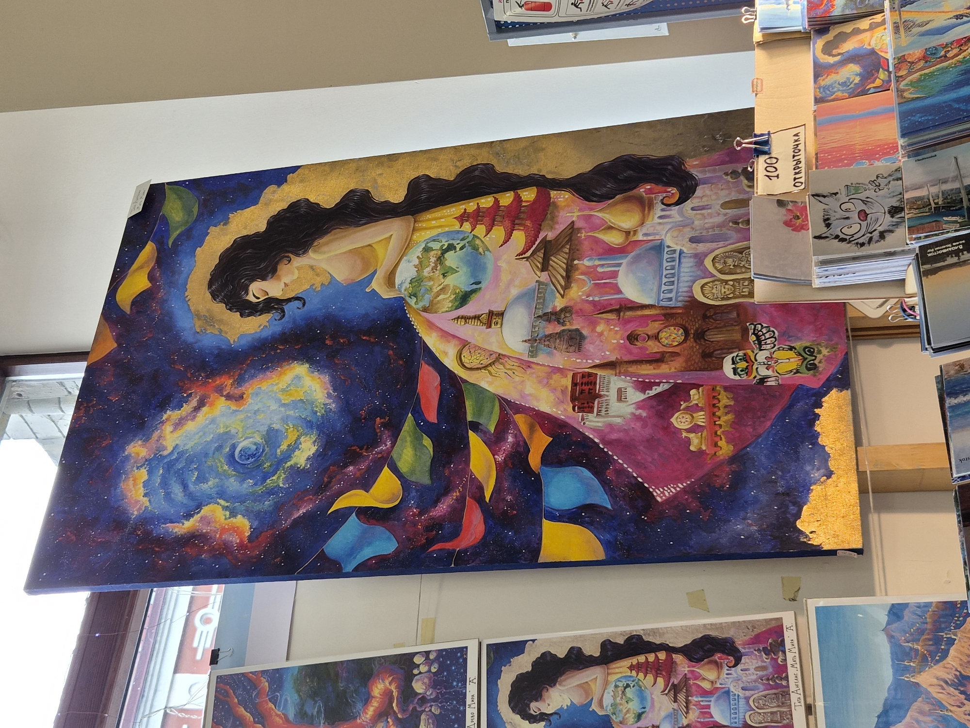 The image displays an art exhibition, likely in Vladivostok, Russia, given the presence of the text "ОТКРЫТОЧКА" (postcards) and Cyrillic script on some of the displayed items. The focal point is a large, vibrant painting of a woman with long, dark hair, adorned with a golden halo. Her dress is a surreal landscape, featuring celestial elements, colorful flags, and a montage of global architectural landmarks, including pagodas, minarets, and domes. The woman holds a representation of Earth in her hands.
To the left of this central artwork, two smaller paintings are visible. The upper one depicts a fiery, tree-like or dragon-like form against a blue and red sky. The lower one shares stylistic similarities with the large central piece, featuring a woman holding the Earth, surrounded by Asian architectural elements.
Below the paintings, a display of postcards and smaller artworks is arranged. One prominent postcard features a stylized black and white cat holding a glass. Another card displays a sunset over a body of water.
The exhibition appears to be set up near a window, with natural light illuminating the scene. The overall impression is one of a vibrant artistic presentation, possibly in a gallery or market setting.