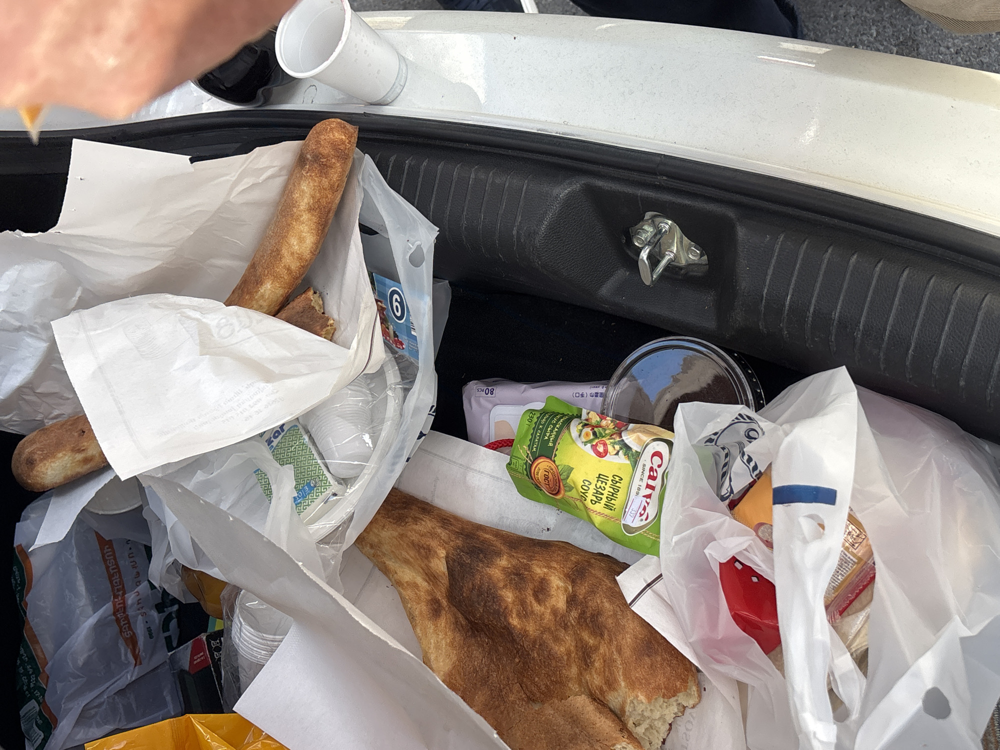 A cluttered car trunk contains an assortment of items including two types of bread, cups, and packaged goods like "Calve сырный цезарь соус" (Caesar cheese sauce) and "nuts". The presence of Armenian text and writing in Cyrillic suggest the location may be in Dilijan, Armenia. A hand is visible on the upper left. The trunk appears to be a white or light-colored vehicle.