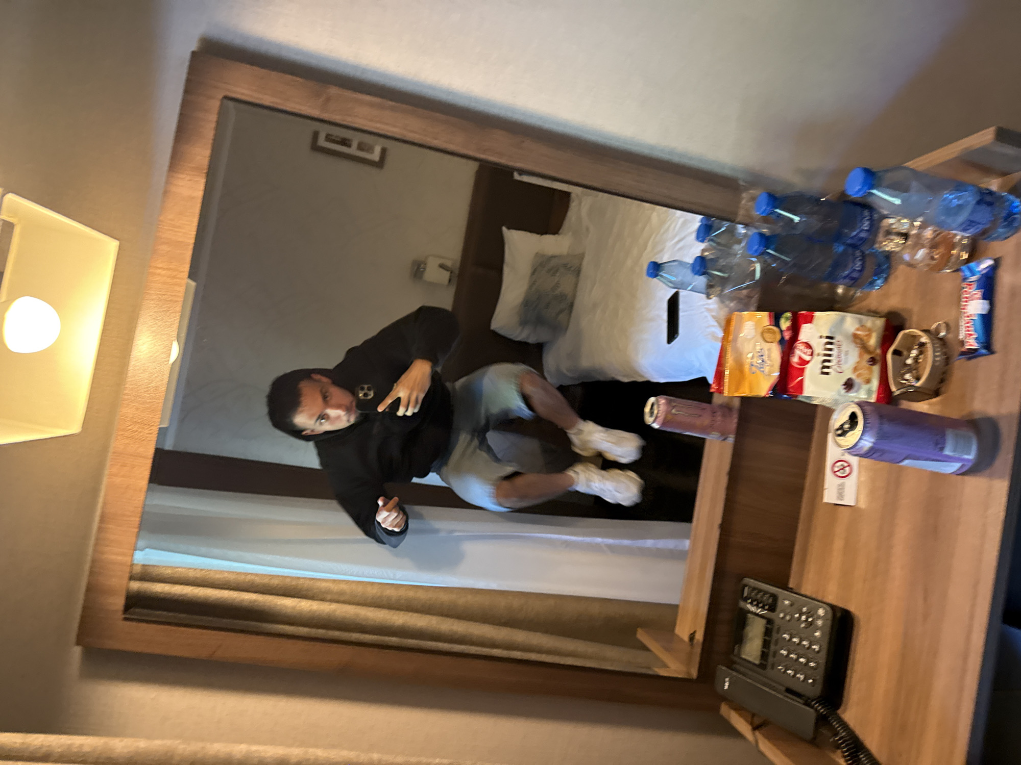 A person is reflected in a hotel room mirror. The person, likely male, is wearing a black hooded sweatshirt and light-colored shorts. They are holding a phone and taking a picture of themselves. The reflection shows a bed with white linens and pillows. A nightstand is visible in the foreground, holding several bottles of water, snack packages, a metal ashtray, a candy bar, an unopened canned drink, and a hotel phone. The room's wall behind the person is painted a light beige color. It is not possible to confirm the location of this image in Veliko Tarnovo, Bulgaria, without further information.