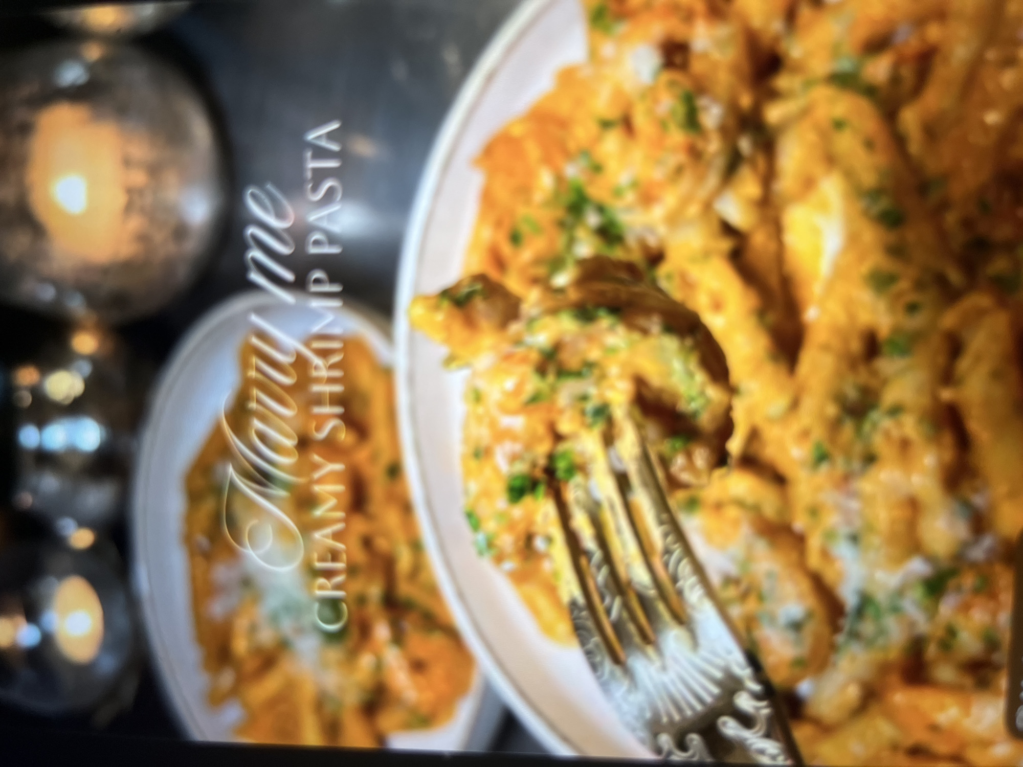 The image shows a close-up of a plate of creamy shrimp pasta. The pasta is coated in a rich, orange sauce and sprinkled with chopped green herbs and white cheese. A decorative silver fork is shown lifting a portion of the pasta, revealing the creamy texture. In the background, another plate of the same pasta is visible, along with decorative candle holders, suggesting a romantic or festive setting. The overall image has a warm and inviting aesthetic. There is no information in the image to suggest a location in Abuja, Nigeria.
