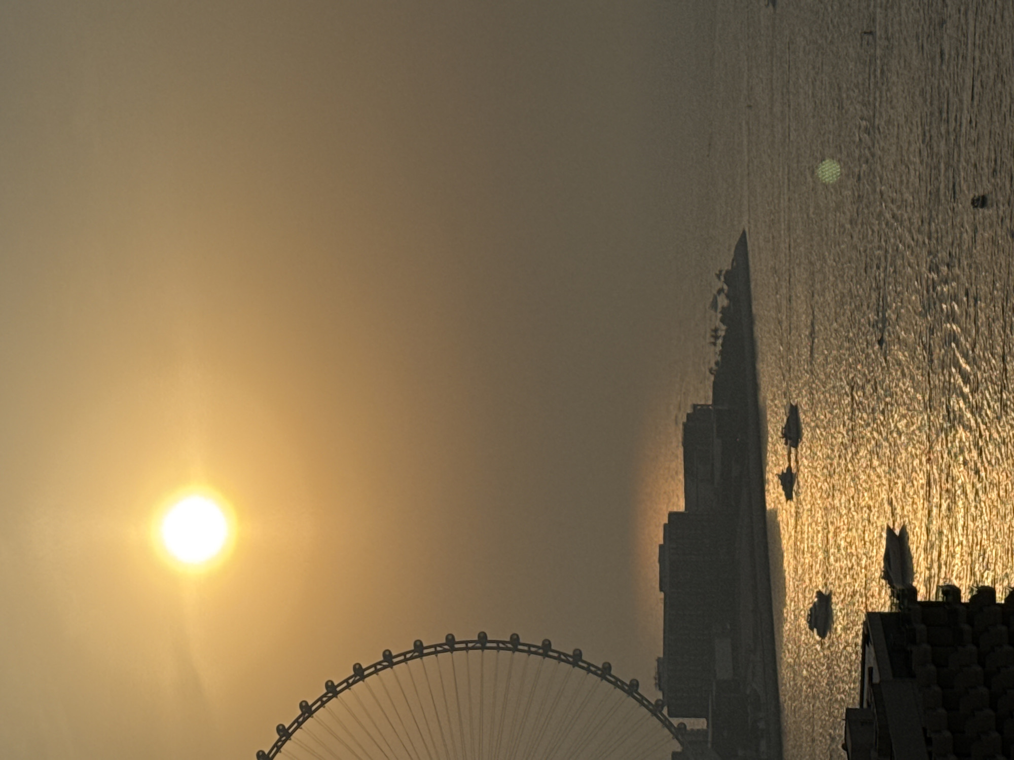 The image depicts a sunset scene over Dubai, United Arab Emirates, likely captured from a high vantage point overlooking the Jumeirah Beach Residence (JBR) area.
In the foreground, the silhouette of a dark, multi-story building is visible on the left, with windows suggesting a residential or hotel structure. The water in the middle ground is illuminated by the setting sun, creating shimmering reflections and ripples. Several boats of varying sizes are present on the water. One boat is moving away from the viewer in the lower center, while a group of three boats are positioned further out in the water to the right.
The middle ground features the skyline of Dubai, characterized by a row of dark, block-like buildings. Behind these buildings, the distinctive structure of the Ain Dubai observation wheel is partially visible on the far left, its massive circular frame silhouetted against the hazy sky.
The background is dominated by a bright, glowing sun low in the sky, casting a warm, orange hue across the scene. The atmosphere appears hazy or dusty, contributing to the soft, diffused light and obscuring distant details. The sea extends to the horizon, meeting the hazy sky.