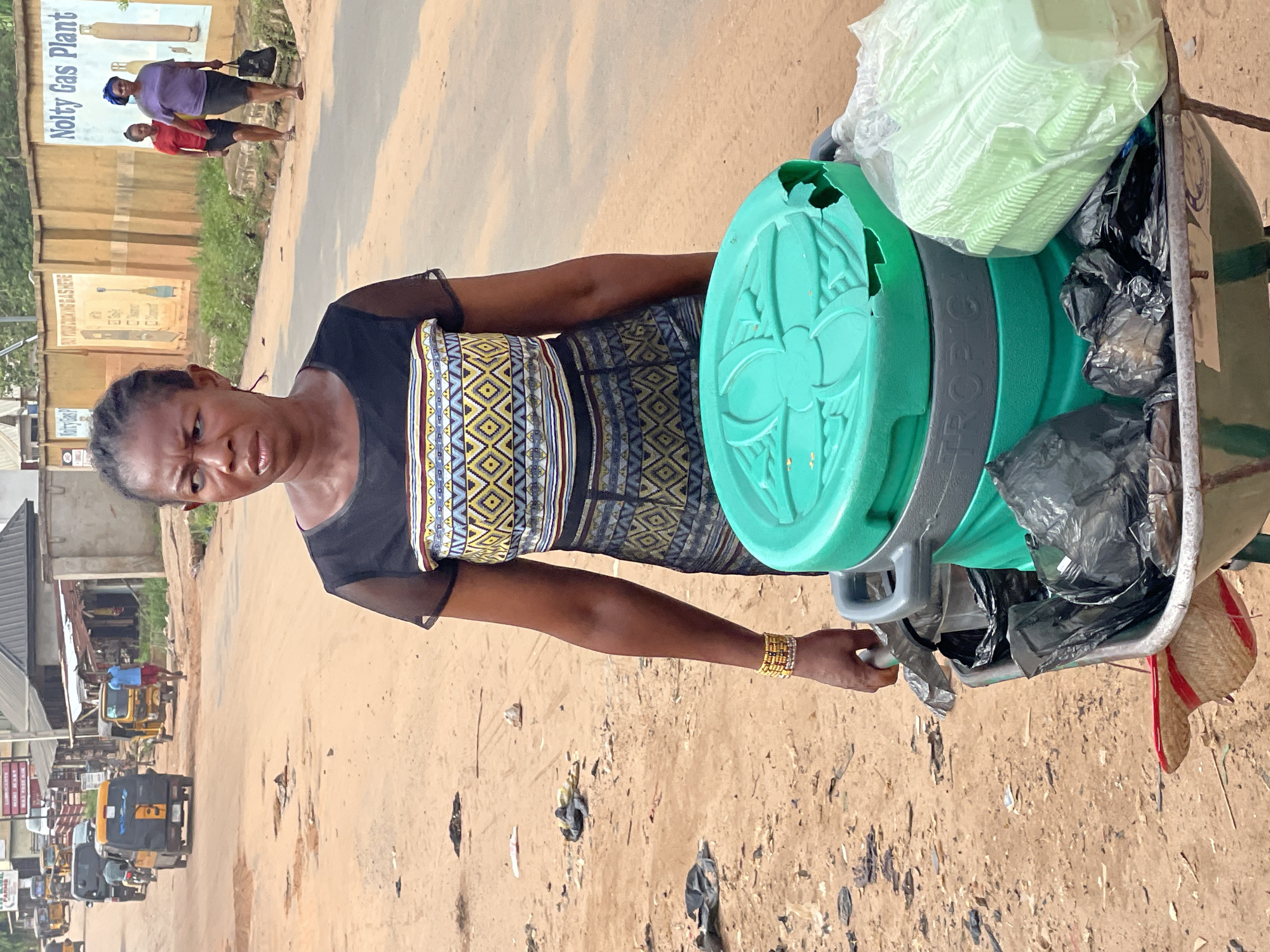 An eye-level outdoor shot depicts a woman in Nkpor, Nigeria, pulling a wheeled cart along a dirt street. The woman is wearing a dark, short-sleeved top with sheer sleeves and a patterned geometric trim at the waist, coupled with a black skirt. She is adorned with several gold-colored bracelets on her left wrist. Her dark hair is styled back into small braids. She is facing the camera with a slight frown. The cart contains green plastic lids, a stack of transparent plastic-wrapped items, and black plastic bags. The ground is littered with debris. In the background, blurred images of shops, signs, and vehicles are visible, including several auto rickshaws in the distance. Other people are visible walking along the side of the road. A sign on the left side of the road reads "Nolty Gas Plant".