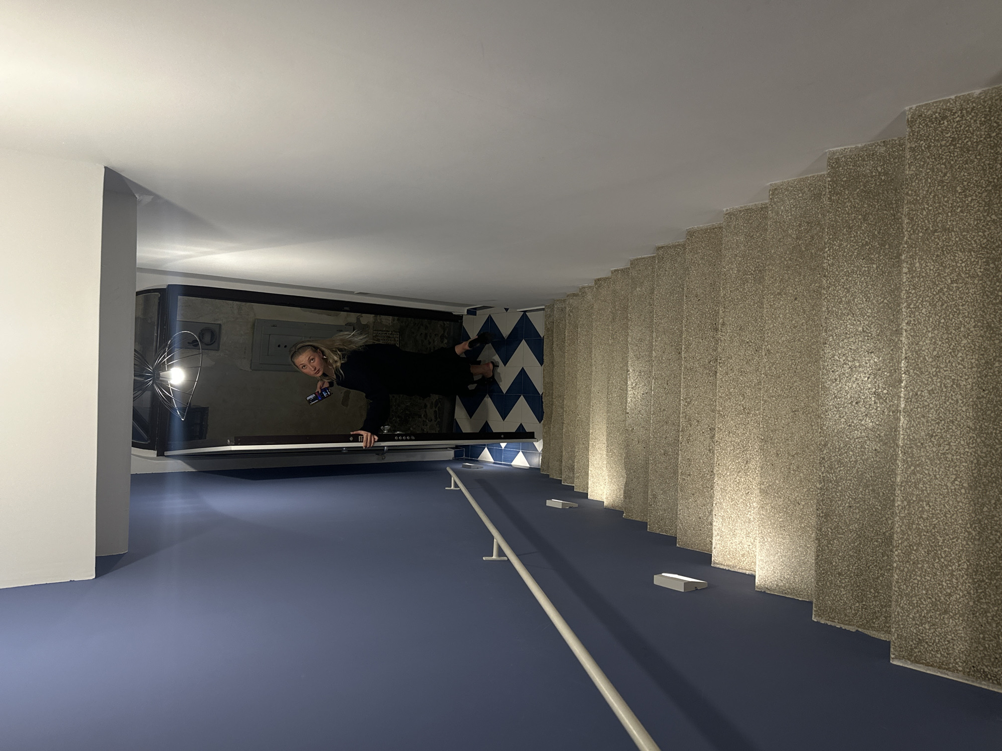 A woman stands upside down in a room, possibly in Lipari, Italy. The room has a blue floor, a white handrail, and a pool table against the wall. A staircase with stone steps runs along one side. The walls are white.