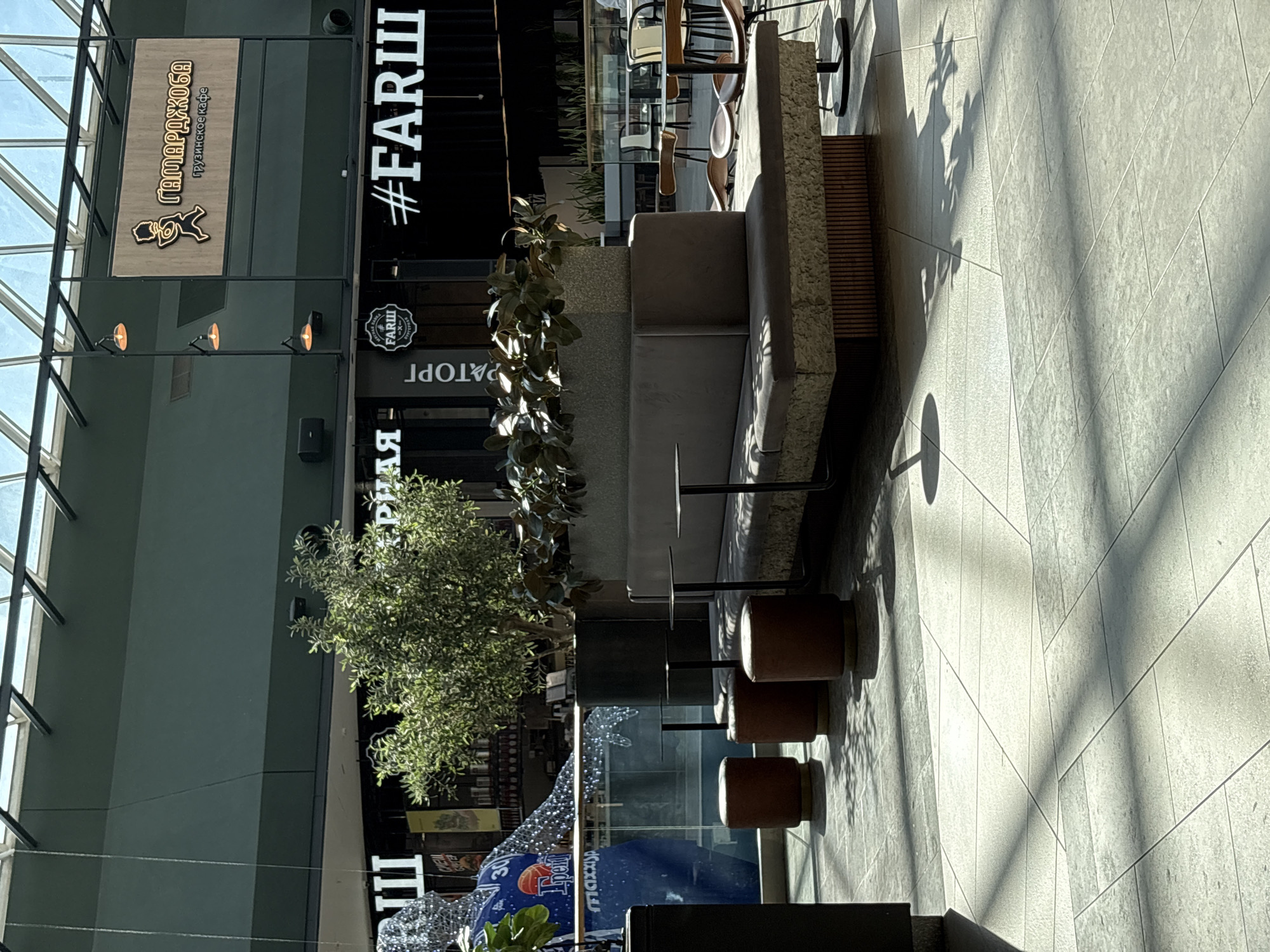 This photograph depicts an interior space in Perm, Russia, likely a shopping mall or commercial building, featuring a seating area.
In the foreground, there is a concrete-textured bench with seating, adjacent to small, wooden cylindrical stools or ottomans. Small tables are placed between the bench and the seating.
Further back, a large tree or plant is situated near a glass wall or window. Beyond the glass, part of a fountain or water feature is visible, along with a decorative installation featuring lights or reflective materials. Storefront signs, including one with the text "#FARSH" and a Georgian restaurant sign "GAMARJOBA," are visible on the left.
The interior space is brightly lit, with sunlight streaming through the glass roof, creating shadows on the tiled floor.