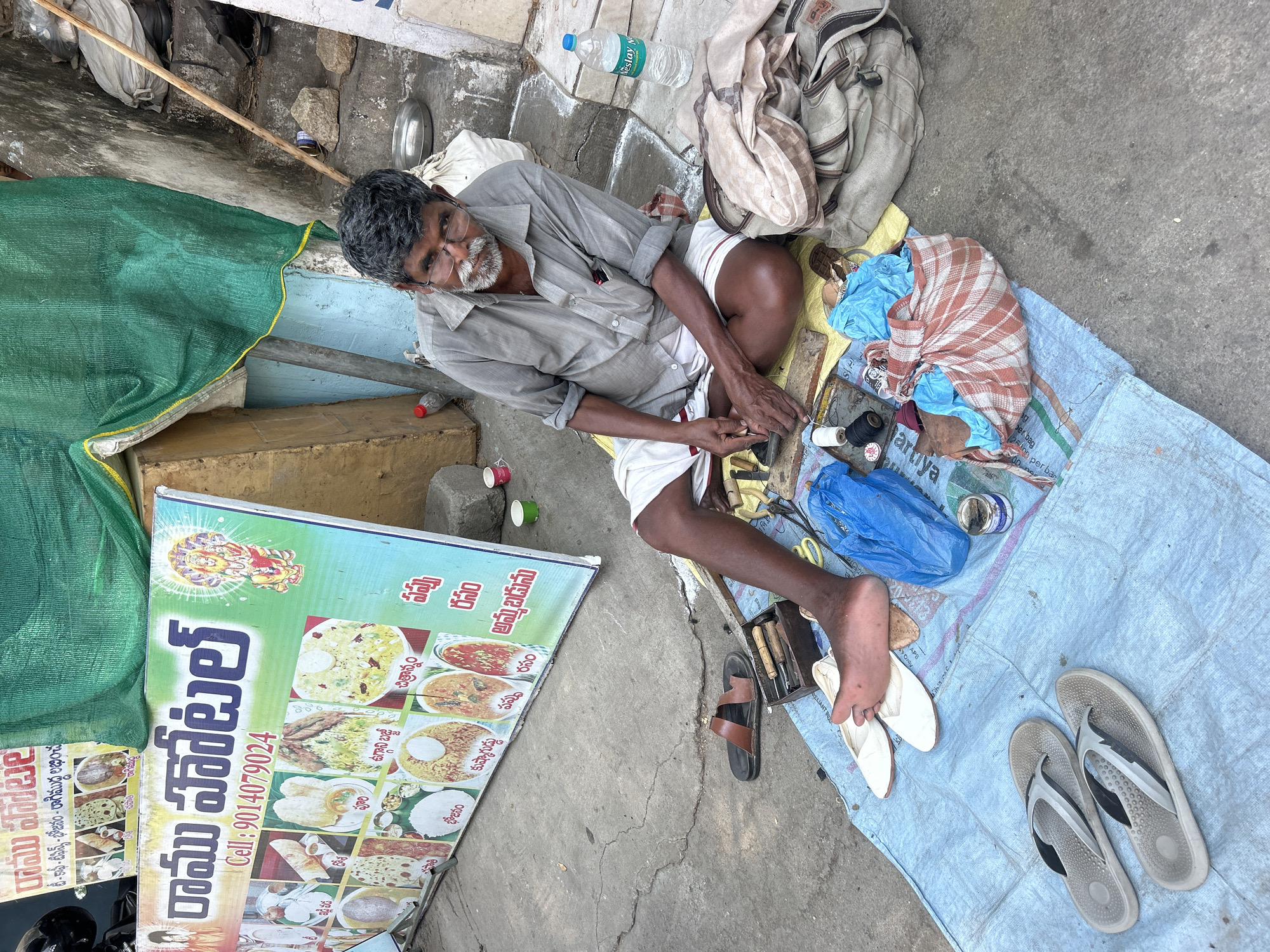 Here is a detailed description of the image:
**Scene:** An outdoor scene in Puttaparthi, India. The image is taken from a high-angle perspective. A man is sitting on a blue plastic sheet spread on the ground, surrounded by tools and supplies, suggesting he is a cobbler or a similar tradesman. A sign for "Sri Ramu Hotel" is propped up on the left side of the frame, with images of food items and a phone number.
**People:** An older man with gray hair and a beard is the central figure. He wears a light gray shirt and white shorts. He is actively working with tools in his hands, seemingly repairing or making shoes.
**Subjects/Objects:**
* **Cobbler's tools:** Various tools, including awls, knives, and possibly a hammer, are scattered around the man. There are also spools of thread and other repair materials.
* **Footwear:** Several pairs of sandals and shoes are present, including the pair the man is likely working on.
* **Plastic sheet:** A blue plastic sheet serves as the man's workspace, providing a surface to work on and keep his tools organized.
* **Signboard:** The "Sri Ramu Hotel" signboard displays images of various South Indian dishes such as idli, vada, poori, and rice meals. It also has the hotel's name in Telugu and English, along with a phone number.
* **Other items:** A water bottle, a folded cloth bag, cups, and other personal items are also visible in the scene.
**Actions/Interactions:** The main action is the man working on shoes, indicating his profession and daily routine.
**Environment:** The scene is likely a street-side or pavement setup, with the man working near the hotel or a similar establishment. The environment appears to be typical of a busy Indian street with exposed concrete and basic amenities.