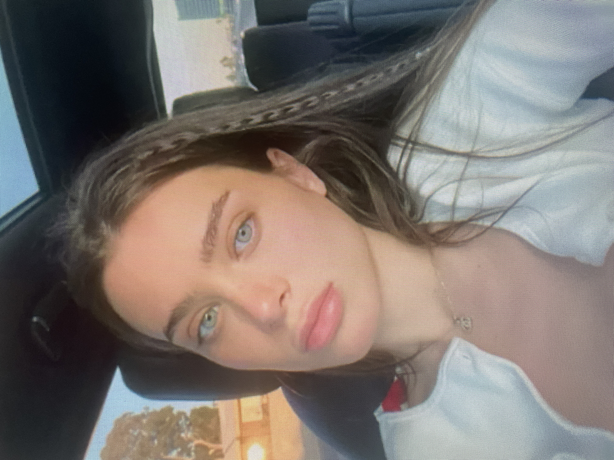A medium shot captures a young woman with blue-green eyes and fair skin, seated in the passenger seat of a car. Her brown hair is styled in braids that fall forward over her shoulders. She is wearing a white cardigan over a red top, and a delicate necklace with a pendant is visible. The background shows the interior of the car, with a sunroof, and hints of an outdoor environment with trees and buildings visible through the windows, suggesting an urban setting at sunset.