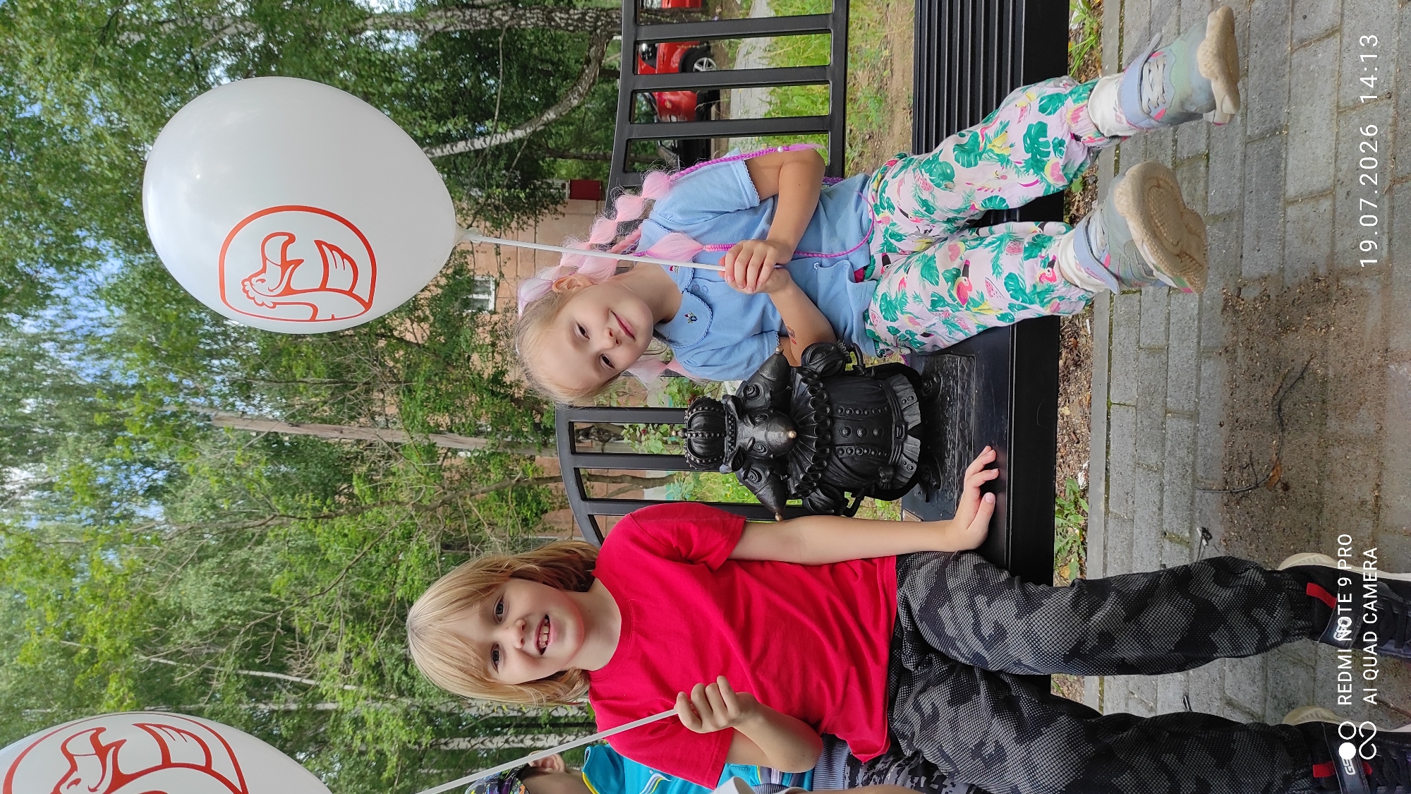 Two young girls, one with blonde hair wearing a red t-shirt and patterned camouflage pants, and the other with blonde hair in pigtails wearing a blue shirt and floral pants, are sitting on a black park bench. The girl on the left holds a white balloon with a red bird logo and a white stick. The girl on the right also holds a white balloon with a similar red bird logo. Between them on the bench is a dark, ornate statue of a figure wearing a crown and ruff. Behind the bench is a line of trees and a building is visible in the background, along with a red car. The foreground shows a paved walkway. The image is dated 19.07.2026 14:13 and includes text overlay indicating "REDMI NOTE 9 PRO AI QUAD CAMERA". The location is identified as Chaikovsky, Russia.