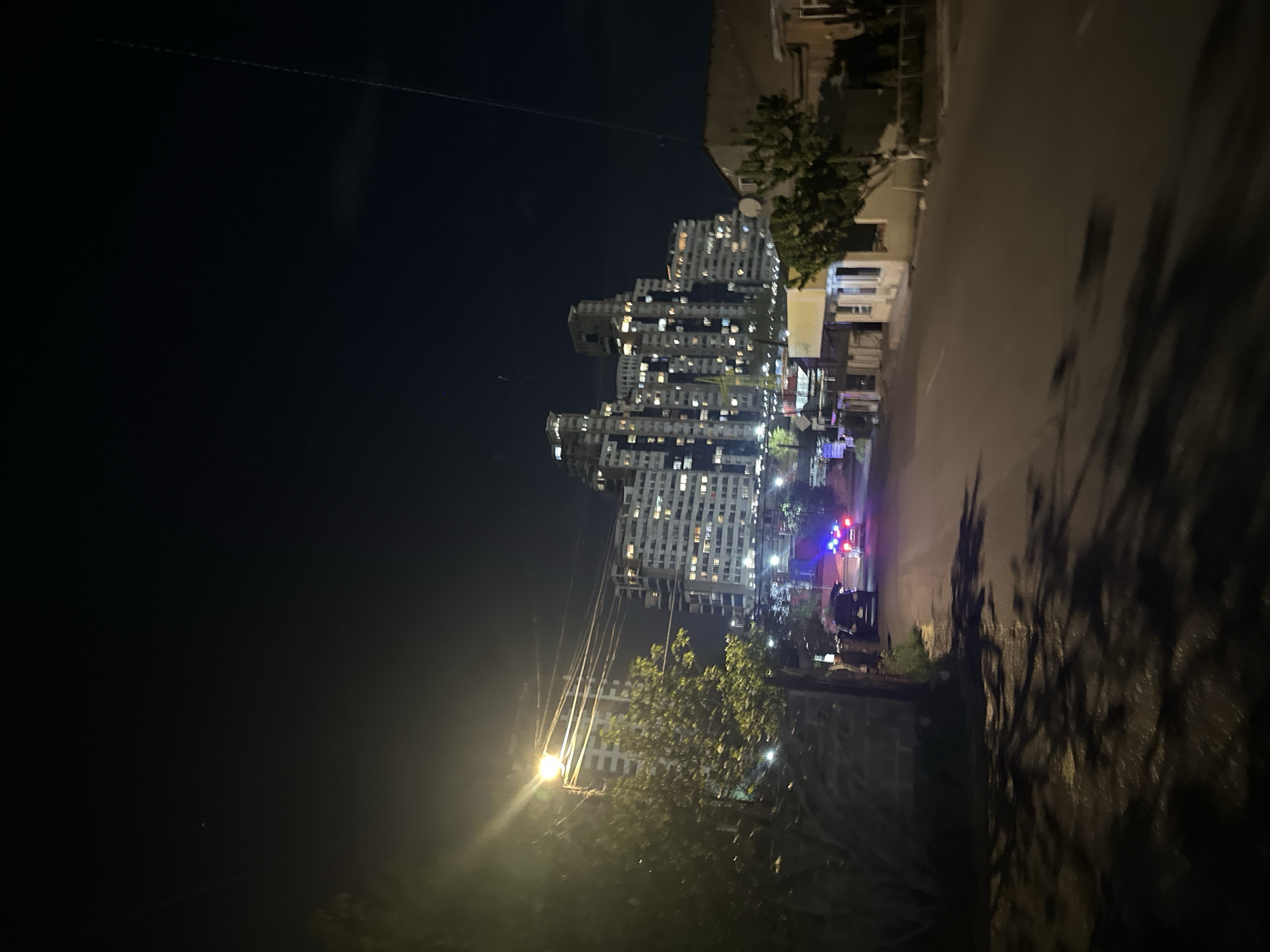 This is a nighttime photograph capturing an urban street scene in Yerevan, Armenia. The image, rotated 90 degrees clockwise from its likely original orientation, prominently features modern high-rise buildings on the left side, characterized by numerous lit windows indicating active residences or offices. Some of these structures appear to have varying architectural styles, with some showing what might be balconies or construction elements.
To the right, a paved road stretches into the distance, illuminated by streetlights and the ambient light from the buildings. On the road, a vehicle with distinct flashing red and blue lights is visible, strongly suggesting the presence of a police car or other emergency service vehicle. Another dark vehicle is parked nearby.
Sparse trees and bushes line parts of the street and foreground, with one particularly prominent streetlight in the lower-middle section casting a bright glow on nearby foliage and power lines stretching across the upper portion of the frame. Long, distorted shadows are cast on the road surface, likely from trees and other structures, adding to the nighttime ambiance. The sky is dark, indicating late hours. No individuals are clearly discernible in the frame, but the active lights and emergency vehicle suggest an ongoing situation or typical urban night activity. There is no visible text.