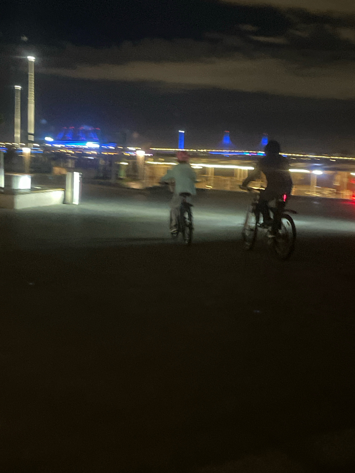 Two individuals are riding bicycles at night along a paved walkway in front of a cityscape. The person on the left is wearing a light-colored shirt, and the person on the right is wearing a darker shirt. The cityscape features illuminated structures, including tall, slender towers with lights at the top, and a larger, dome-shaped building with blue and red lights. The sky is dark with visible clouds. The image appears to be taken at night in an urban environment, possibly near a waterfront. The location is the area around the Lakhta Center in Saint Petersburg, Russia, which is known for its distinctive architecture and waterfront promenade.