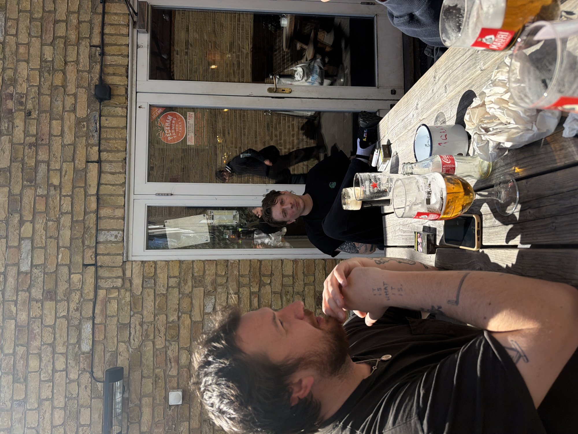 Here is a description of the image:
The image shows a group of people sitting at an outdoor table next to a building. There are at least three people visible, one of whom is in the foreground with tattoos on his arm. There are numerous empty beer glasses and bottles on the table, along with a pack of cigarettes and a mobile phone. A sticker on the door behind the table indicates that the location is the "Peckham Farmers Market" in the SE15 postcode of London. The building appears to be made of brick and has a white door and window frames. There are other people visible through the windows of the door. The general setting suggests an outdoor seating area of a pub or restaurant.