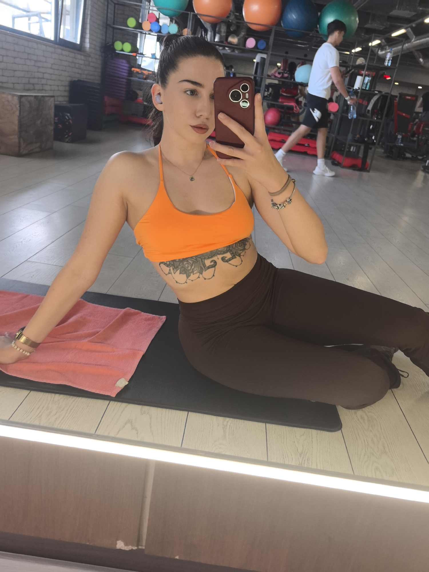 The image shows a young woman indoors, seemingly at a gym. She is fair-skinned, with dark hair pulled back, wearing an orange sports bra and brown leggings. She has a tattoo on her abdomen and bracelets on her wrists. She is holding a phone up, as if taking a selfie. She is sitting on a black exercise mat with a pink towel beside her. In the background, there is gym equipment and another person walking in a white t-shirt and black shorts. The gym has light-colored floors and is well-lit. The location is most likely in Stara Zagora, Bulgaria, given the prompt.