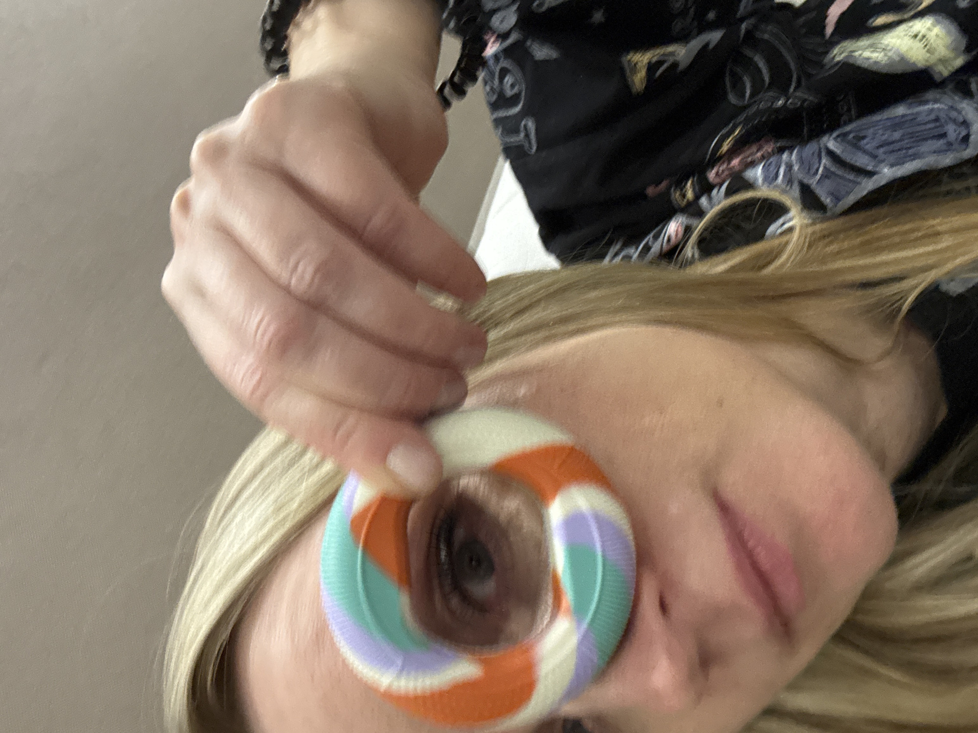 A close-up, angled image captures a subject, identified as a woman with blonde hair, centered in the frame. Her right eye is visible through the central aperture of a textured, ring-shaped object held by her left hand. The object displays a distinct spiral pattern in orange, light green, and light purple. Additional facial features visible include her nose, mouth with slightly parted lips, and chin. Her left wrist bears a dark, possibly beaded bracelet. The woman is wearing a dark-colored garment with an abstract, multi-tonal pattern visible on the right side of the frame. The background consists of a plain, light-colored interior surface, largely out of focus. This scene is situated in the city of Votkinsk, Russia.