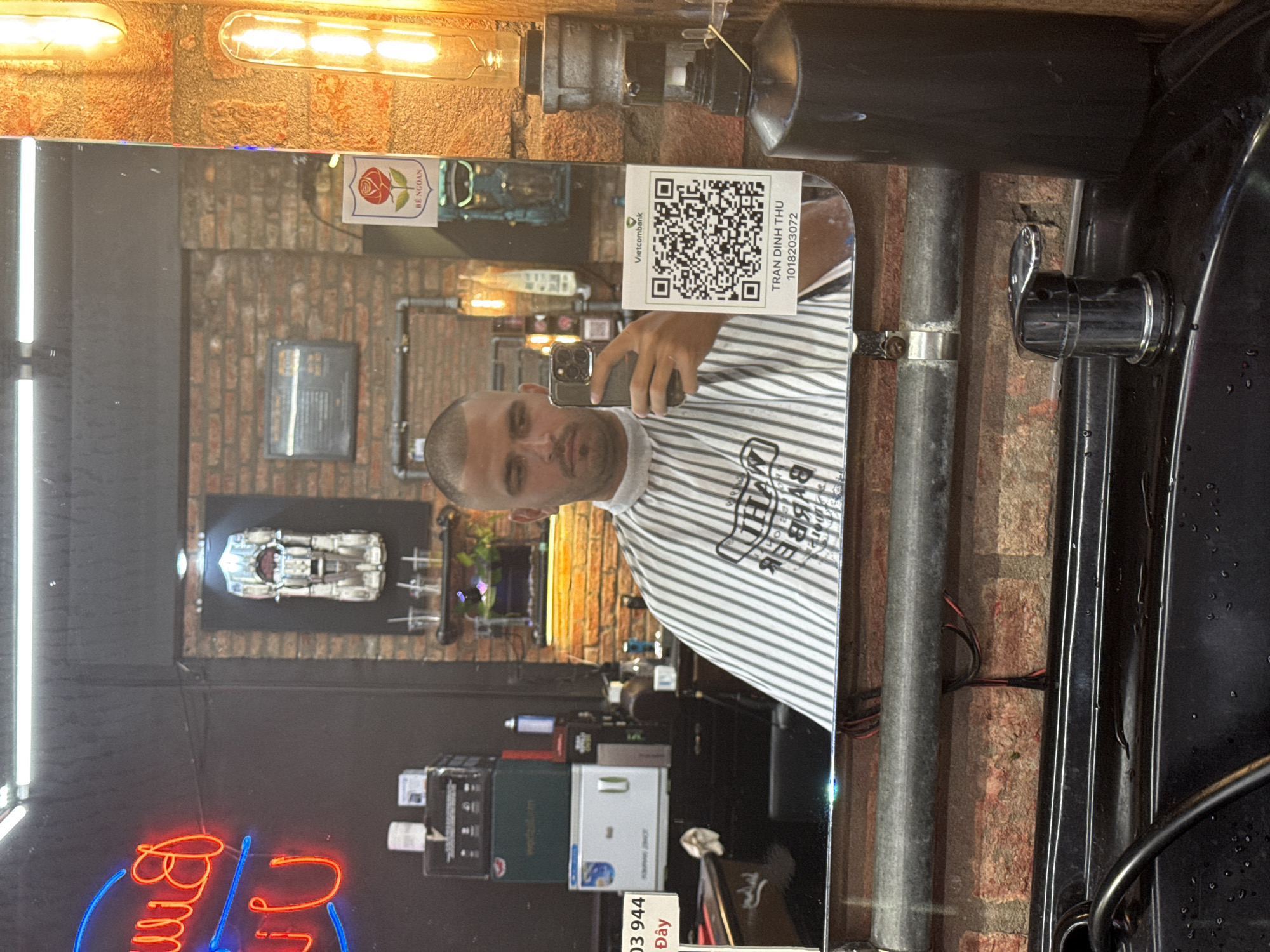 A man takes a selfie in a mirror at a barber shop. The mirror is mounted on a brick wall with exposed pipes. The shop features neon lighting, a model car display, and a variety of hair care products. A QR code sticker is visible on the mirror with the name "TRAN DINH THU" and account number "1018203072." Vietnamese text is present in the image. The location is likely Đà Nẵng, Vietnam.