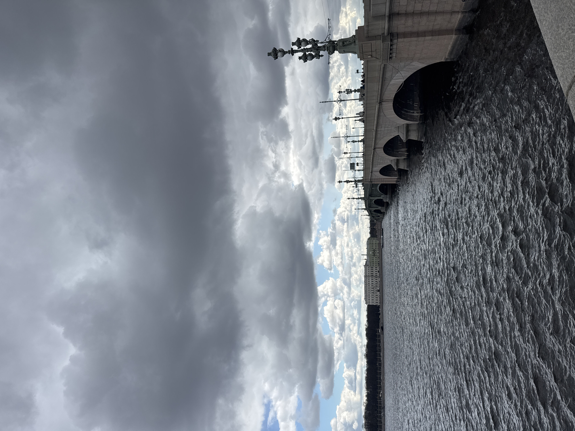 A landscape-oriented, eye-level photograph captures a section of the Palace Bridge over the Neva River in Saint Petersburg, Russia, on a cloudy day.
The right side of the image features the Neva River, its surface ruffled by ripples, rendered in shades of gray due to the overcast conditions. A dark strip of land, possibly a park or the riverbank, runs along the river's edge, and a few buildings are visible in the distance.
On the left, the focus shifts to the sky, dominated by heavy, gray storm clouds. Patches of lighter, brighter clouds are visible, suggesting a clearing or a break in the weather.
The Palace Bridge cuts across the image, its stone arches extending over the river. Ornate lampposts with multiple lamps are mounted along the bridge's edge, contributing to the architectural detail. The bridge’s structure appears to be made of stone or concrete.