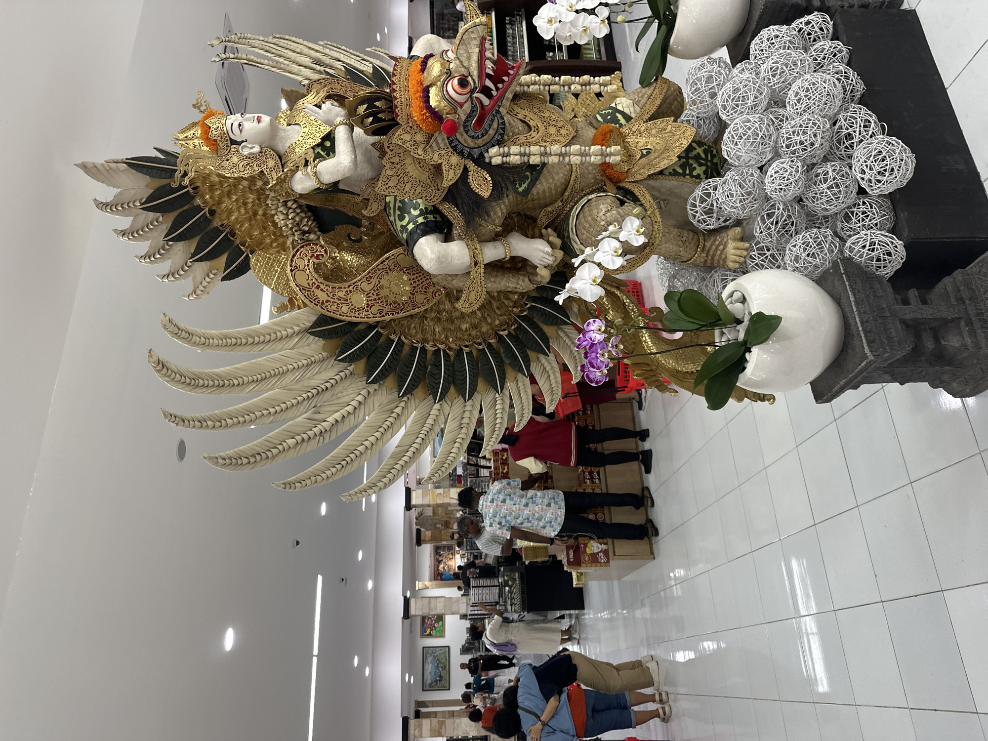This image depicts a large, ornate statue of Garuda Wisnu Kencana in what appears to be an interior location in Kuta, Indonesia. The statue is predominantly gold, with black and white feathers and red and black accents. It is positioned above a retail space, where shoppers browse items displayed on shelves. The floor is tiled and white. The lighting is bright, suggesting a modern or upscale retail environment. The statue is intricately detailed and seems to be a centerpiece or significant decorative element within the space. People in casual clothing are walking around the base of the sculpture. There is a collection of decorative white spheres near the statue. The Garuda figure carries the Wisnu figure on its shoulders.