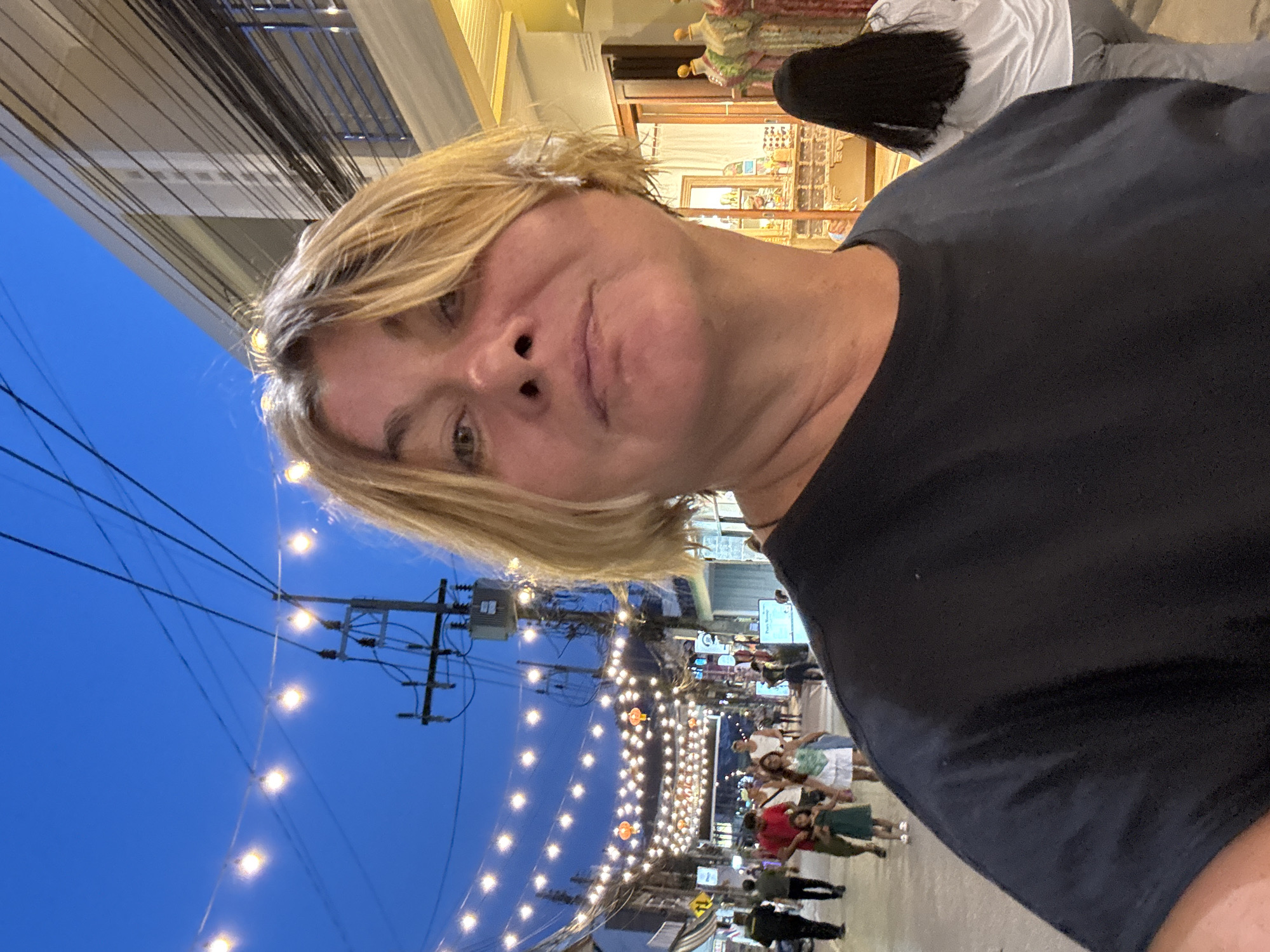 An outdoor selfie is taken at dusk in Baan Bo Phut, Thailand. The foreground features a woman with short, blonde hair wearing a black t-shirt. Her face is partially visible and turned towards the camera. Behind her, a street is lined with buildings and illuminated by strings of fairy lights and some decorative lanterns. Pedestrians are walking along the street, some appearing to be shopping or strolling. The sky above is a deep blue, characteristic of twilight. The environment suggests a lively market or shopping area.