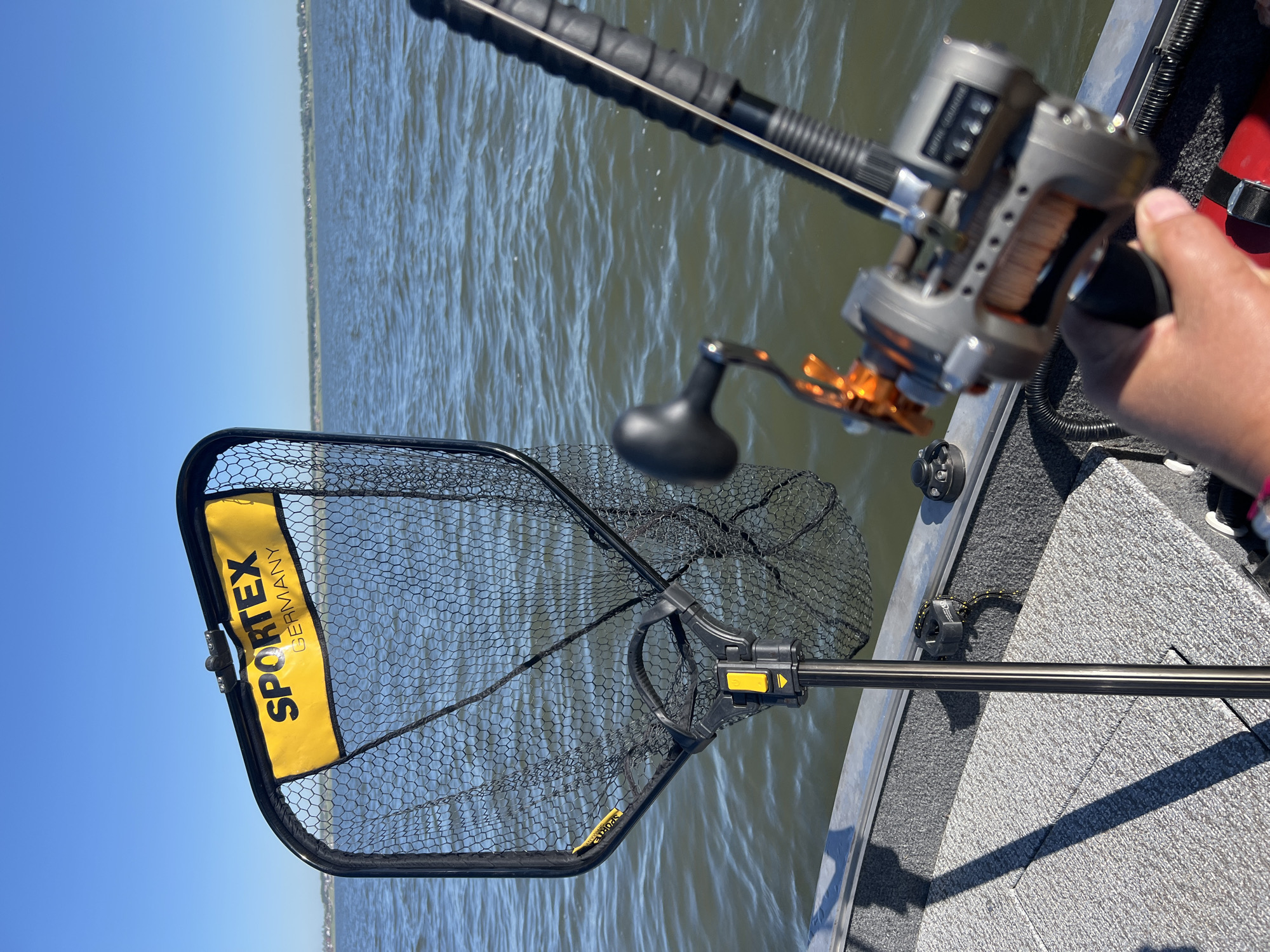 A person's hand holds a fishing reel on a boat. The reel is gray and silver with orange accents and a black handle. A fishing rod extends from the reel. In the background, a fishing net with a yellow banner that reads "SPORTEX GERMANY" is attached to a pole. The boat is on a body of water, likely a lake or river, under a clear blue sky. The far shore is visible in the distance, lined with trees and some buildings. This scene is not located in Novopichugovo, Russia, as there is no information in the image to suggest this location.