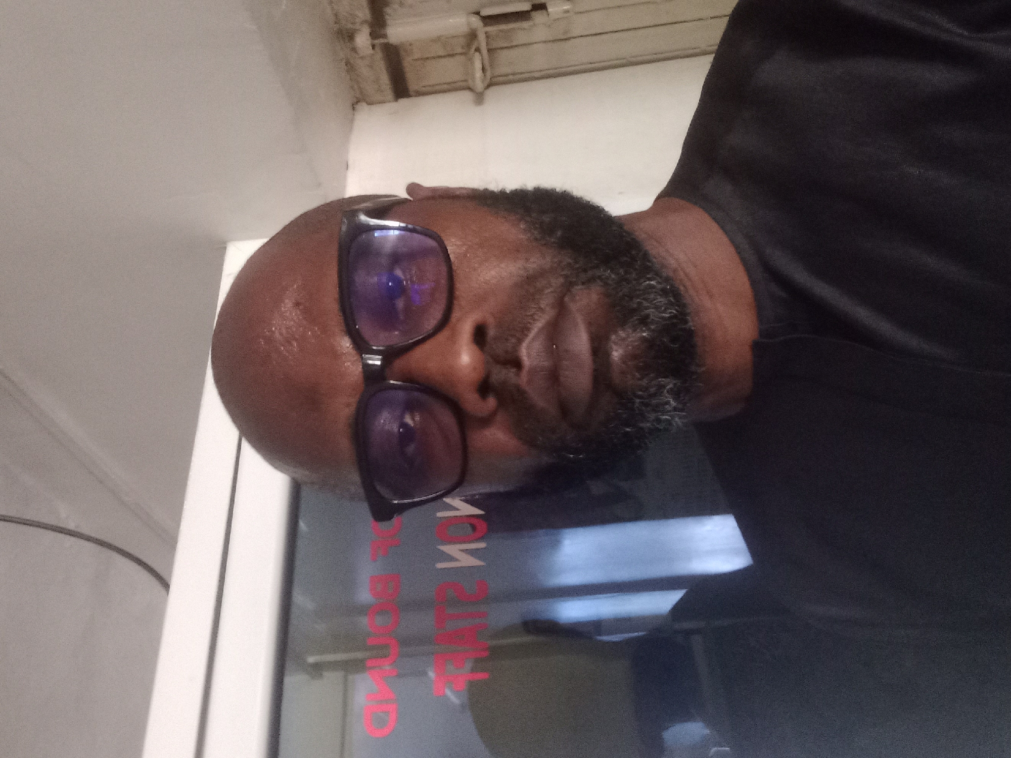 A person with a bald head, beard, and glasses is seen in a close-up, angled view. The person is wearing a black shirt. The background appears to be a window with a red inscription, possibly a reversed or reflective message, and an interior setting. The location and city are Unknown.