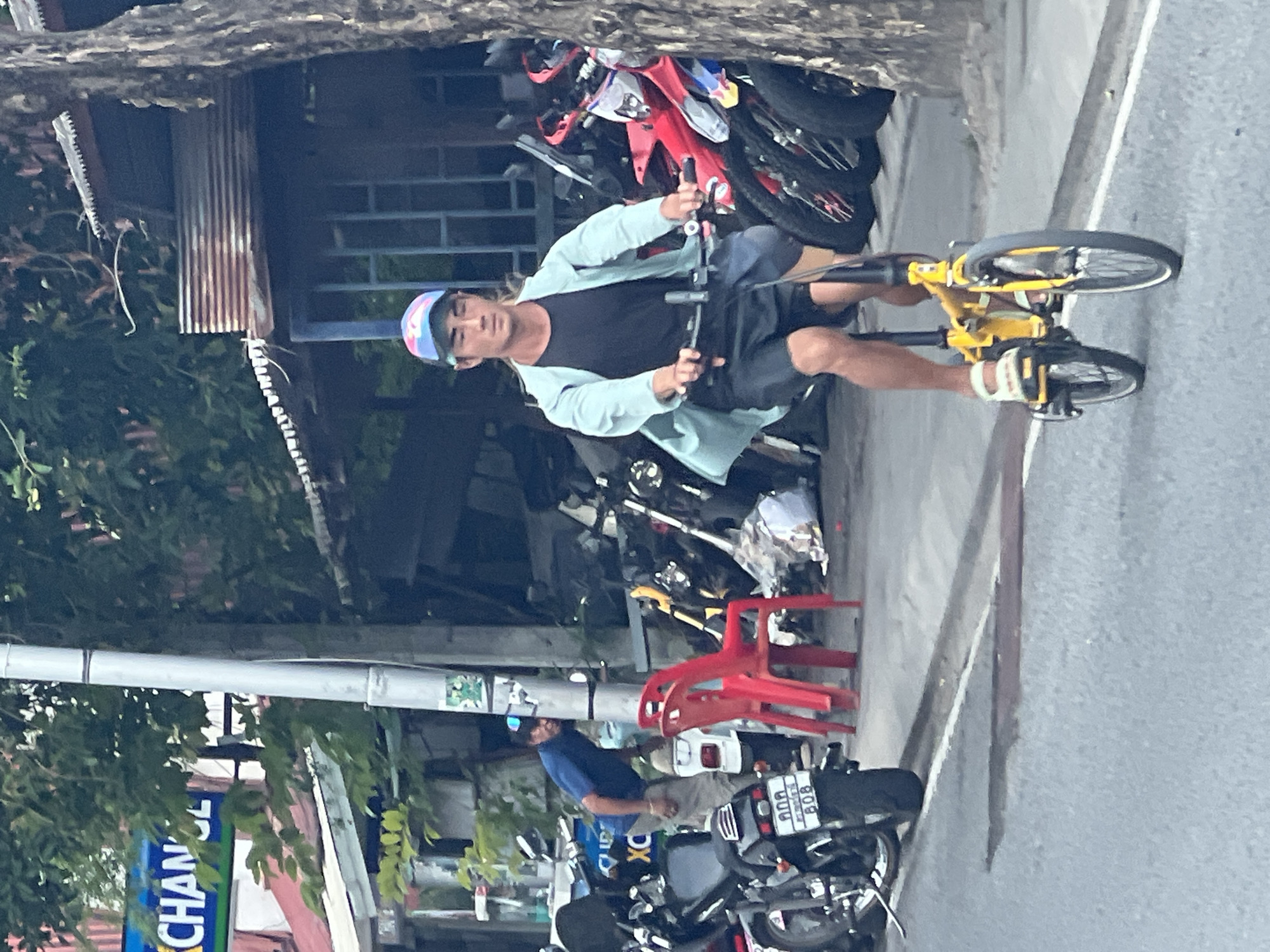 A young East Asian male is riding a yellow folding bicycle down a paved street in Ban Rang Rak, Thailand. He wears a blue and pink baseball cap, a light blue open-collared shirt over a black t-shirt, and dark shorts. His feet are clad in white sandals. He is riding in the foreground, facing forward and slightly left. Behind him, parked on the side of the road, is a collection of motorcycles and scooters, some are red and white. To the left of the motorcycles, a man wearing a blue shirt and beige pants is bent over, looking towards the ground near a motorcycle. Further left, a blue sign with yellow lettering is partially visible, appearing to read "EXCHANGE". Two red plastic chairs are positioned near the parked motorcycles. A utility pole stands to the left of the man in the blue shirt, adorned with stickers. The background shows a building with a corrugated metal roof and a tree trunk on the far right. The street appears to be lined with businesses.