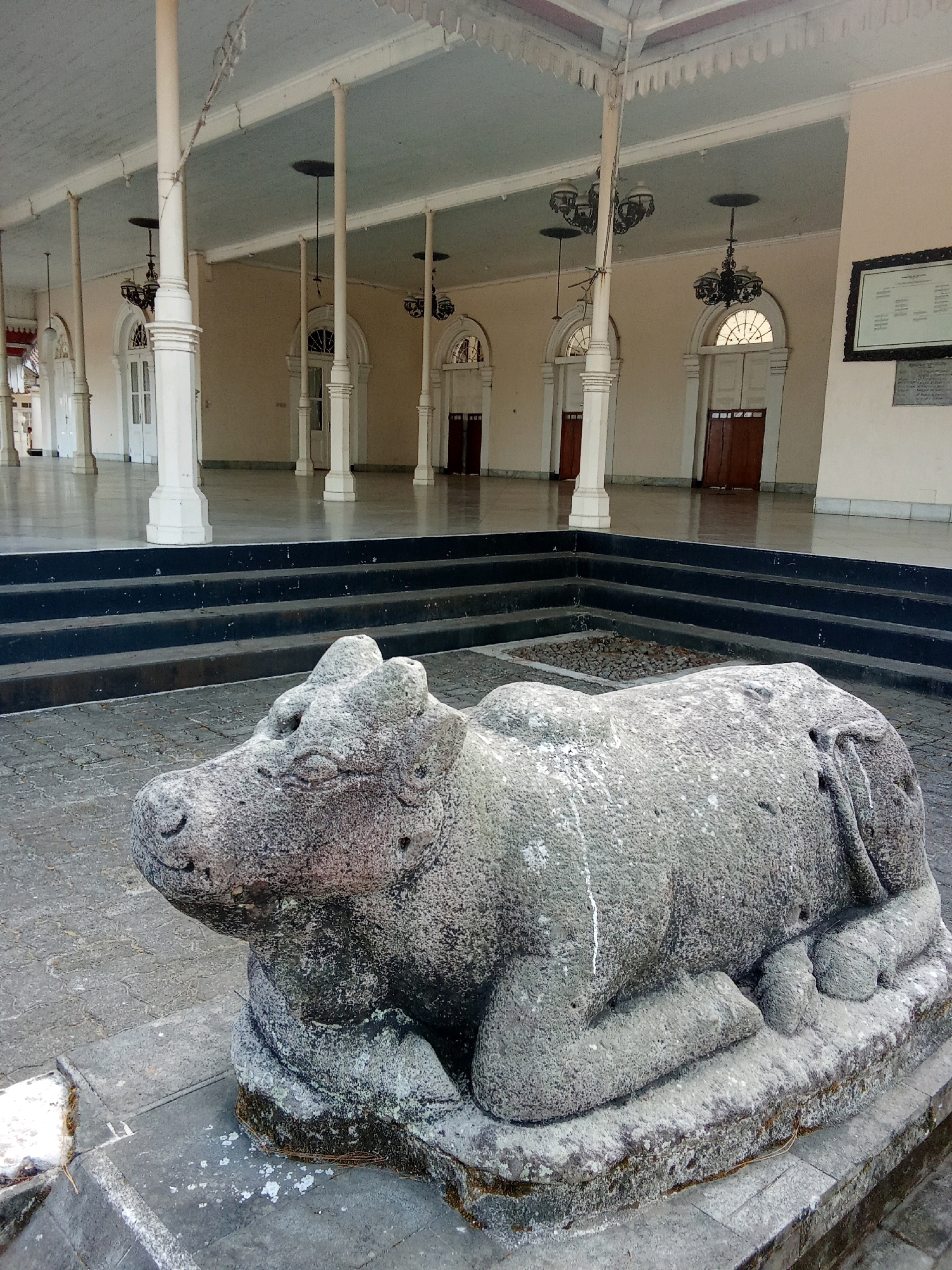 This image depicts a stone statue of a cow, likely representing Nandi, the bull vahana of the Hindu deity Shiva, situated in front of a colonial-era building. The statue is positioned on a paved courtyard, with a dark, multi-tiered platform rising behind it. Beyond the platform, the building features a covered walkway supported by numerous white pillars. The walkway leads to several arched doorways with double wooden doors and ornate windows. Chandeliers hang from the ceiling of the walkway. A framed document is affixed to the building's wall on the right side of the image. The background suggests an open, public space within a historical setting. This scene is located at the complex of the so-called "Rumah Tidar" or "Borobudur Area Gate" in Magelang, Indonesia. The "Rumah Tidar" is an area that serves as a gateway to the Borobudur temple and is known for its historical significance and architectural elements dating back to the Dutch colonial period.