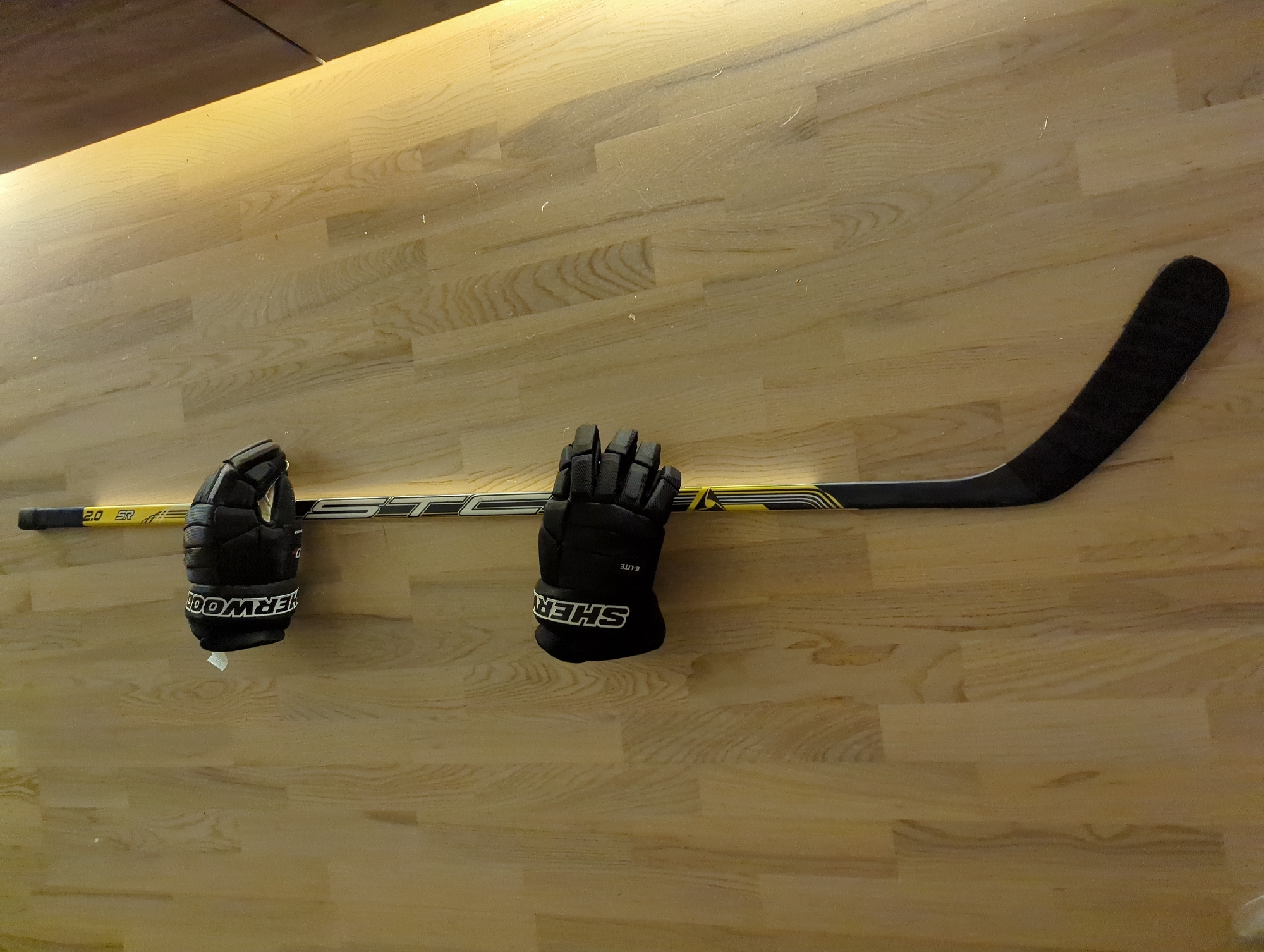 The image shows a hockey stick and a pair of hockey gloves lying horizontally on a light-colored, wooden surface. The hockey stick is black and yellow and is labeled "STC." The gloves are black and have the word "SHERWOOD" printed on them. The background is a light-colored, wooden surface. There is no information present to confirm a location in Minsk, Belarus.