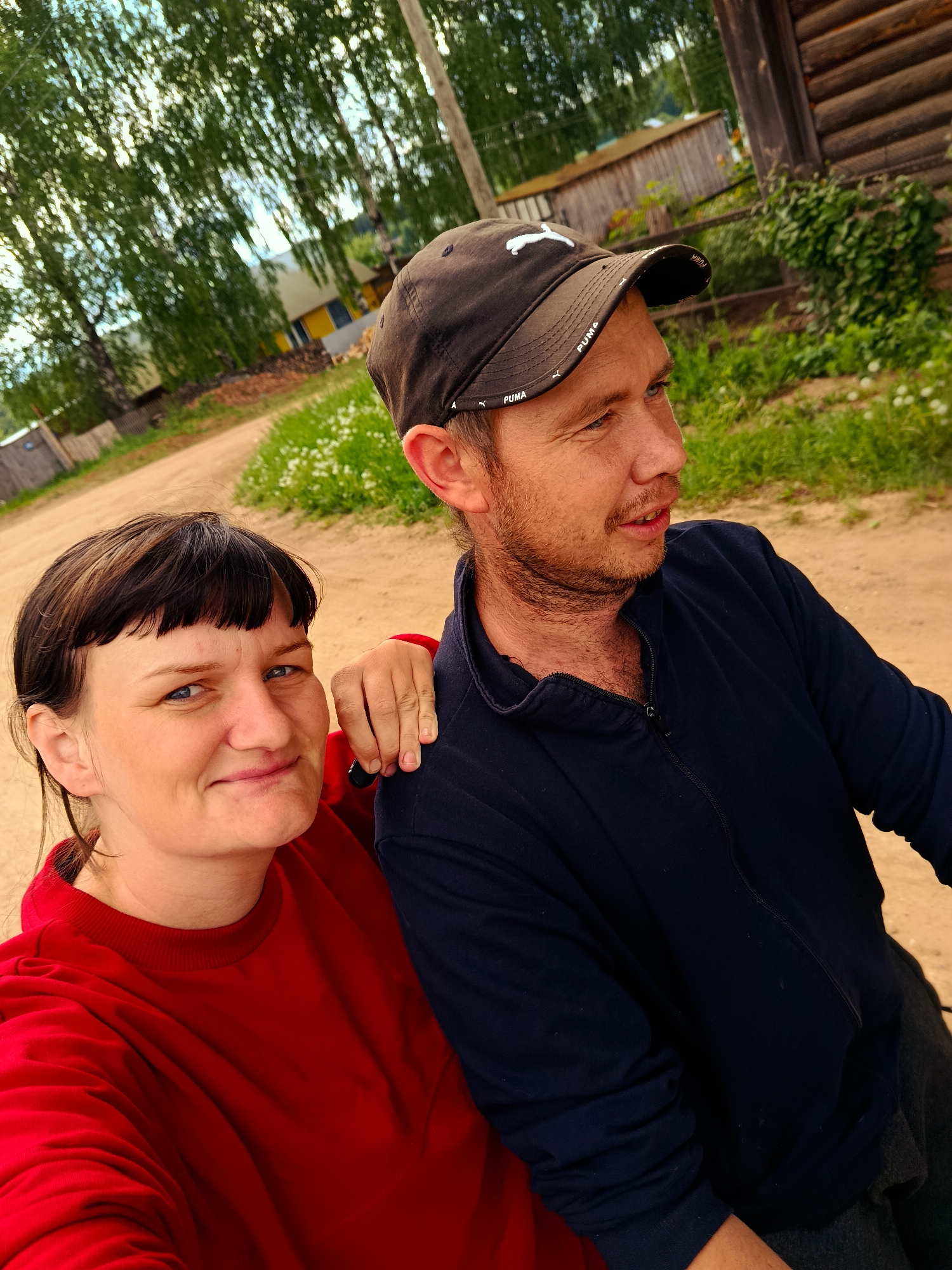 A medium outdoor shot captures a man and a woman on what appears to be a dirt road. The woman, positioned to the left, is wearing a red sweater and has dark hair. Her arm rests on the man's shoulder. The man, to the right, is wearing a black zip-up jacket and a black Puma baseball cap. He appears to be speaking. The background features a dirt road, trees, and a wooden building. The location is Фоки, Russia.