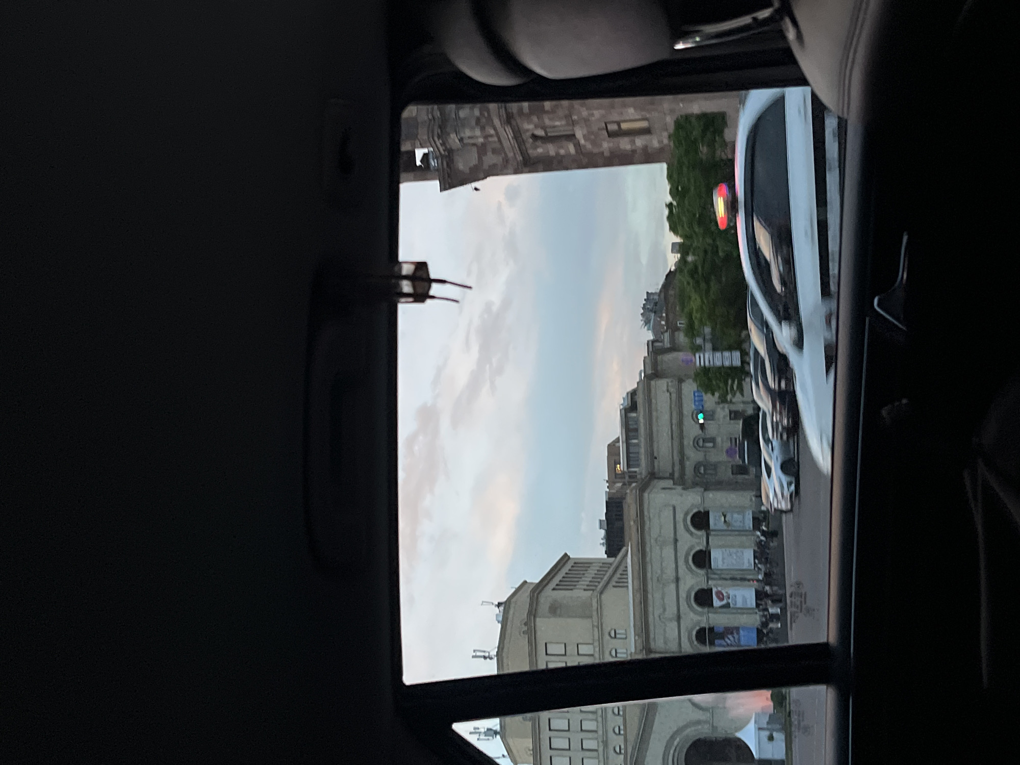 The image captures a view from inside a car, looking out through a window. The scene outside the window features urban architecture with white buildings, indicative of a city setting. The sky is visible, with clouds suggesting it could be dusk or dawn. Several cars are parked or moving along the street. The architecture and general ambiance suggest the image was taken in Yerevan, Armenia, with buildings resembling those in the city's central districts.