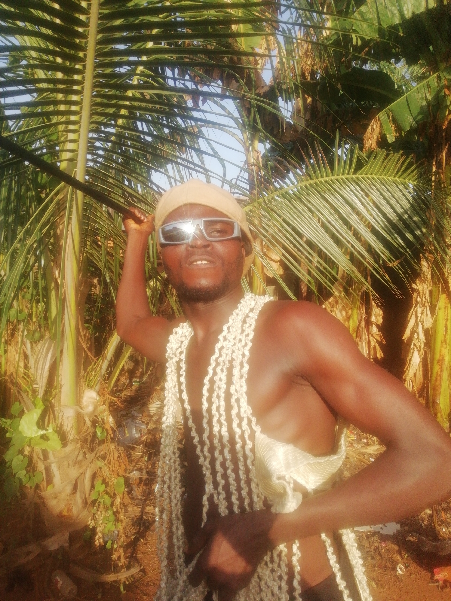 A man is captured from a low-angle perspective in an outdoor scene. He is dark-skinned, wearing a tan cap and reflective sunglasses, and is draped with strings of white shells. He holds a long, dark rod in his raised right hand. The background is filled with palm trees and banana plants, suggesting a tropical environment. The scene is bathed in bright sunlight. The location appears to be a natural setting, possibly in the city of Unknown, Unknown.