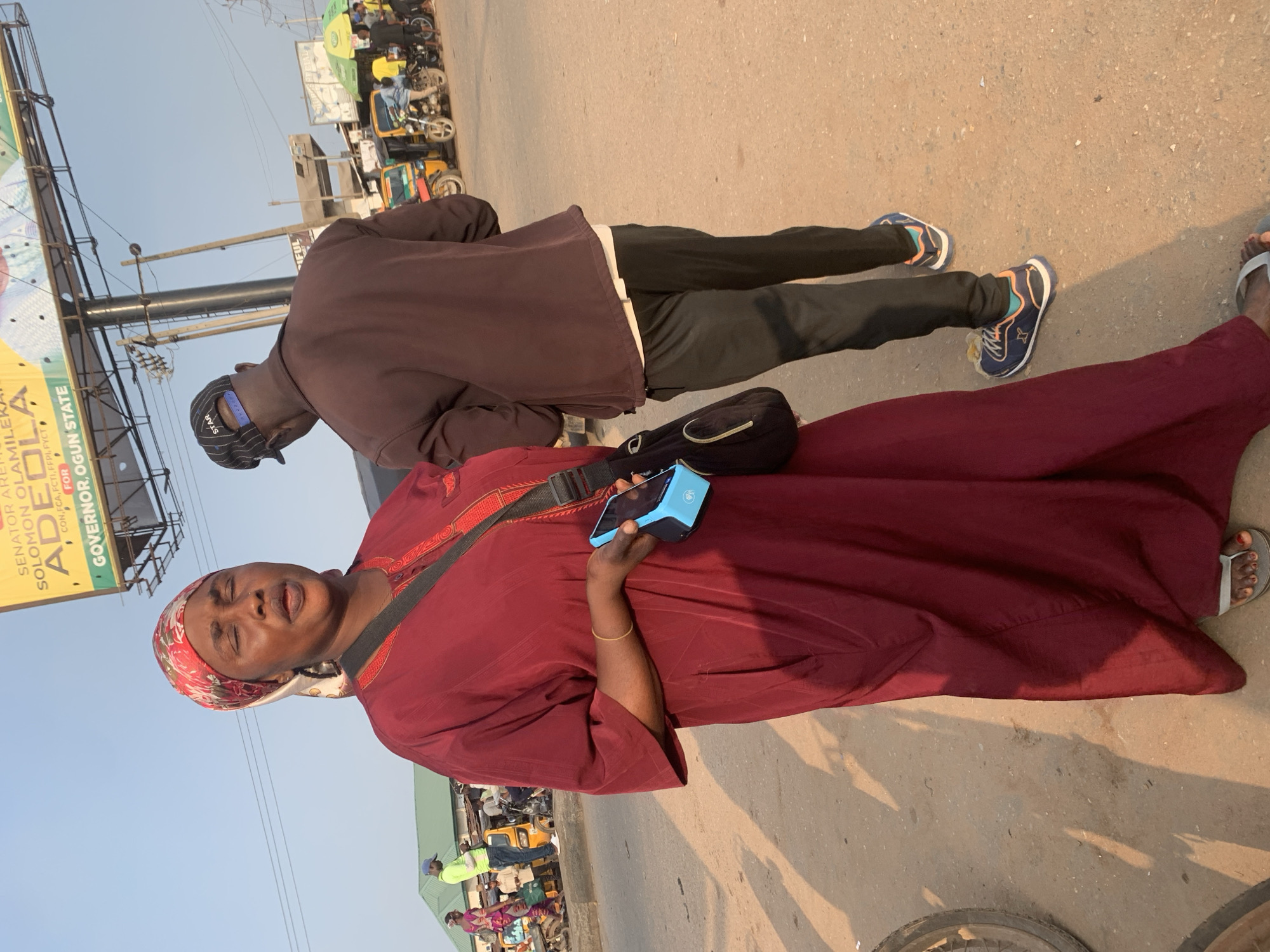 In Shagamu, Nigeria, a woman in a burgundy abaya and headscarf stands with her eyes closed and mouth open, holding a blue card reader. A man in a brown jacket and black pants walks away from the camera in the background. The scene is on a dusty street, with a billboard for "SENATOR ABBA HAMILEKAN" and "GOVERNOR, OGUN STATE" visible overhead. Several vehicles, including a yellow tricycle and motorcycles, are parked along the side of the road. The lighting suggests it is late afternoon.