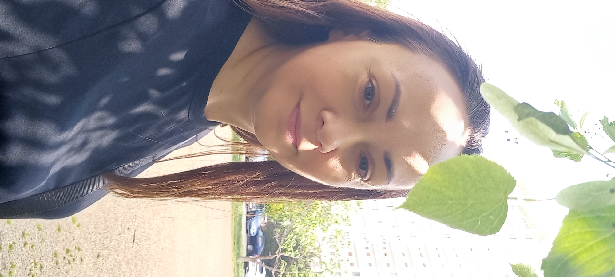 A woman with light skin, long brown hair, and blue eyes looks at the camera. She is wearing a dark blue shirt. Sunlight illuminates her face, and she is standing outdoors near a tree with green leaves. In the background, there is an urban landscape with buildings and cars, suggesting the location is Krasnodar, Russia.