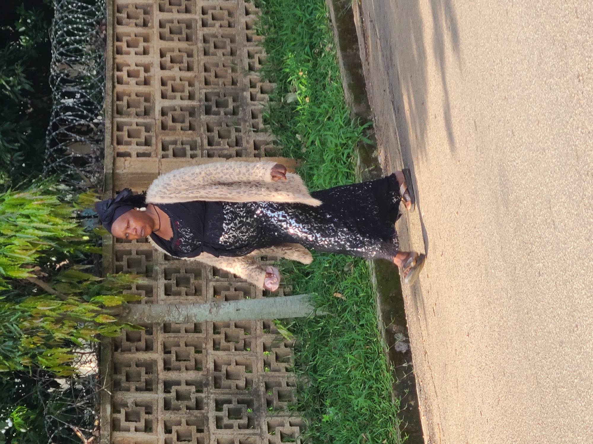 A woman stands outdoors in what appears to be Jos, Nigeria. She is wearing a black sequined dress and a fuzzy, leopard-print coat. The woman has a dark headscarf and is wearing earrings. She is also wearing sandals. The background consists of a concrete block wall with an opening pattern, green grass, and some barbed wire. The ground in front of her is a paved surface, possibly a road or sidewalk. Sunlight is casting shadows on the ground, suggesting it is either morning or late afternoon. The woman's mouth is open, and she appears to be yawning. The overall impression is a casual, everyday moment captured outdoors.