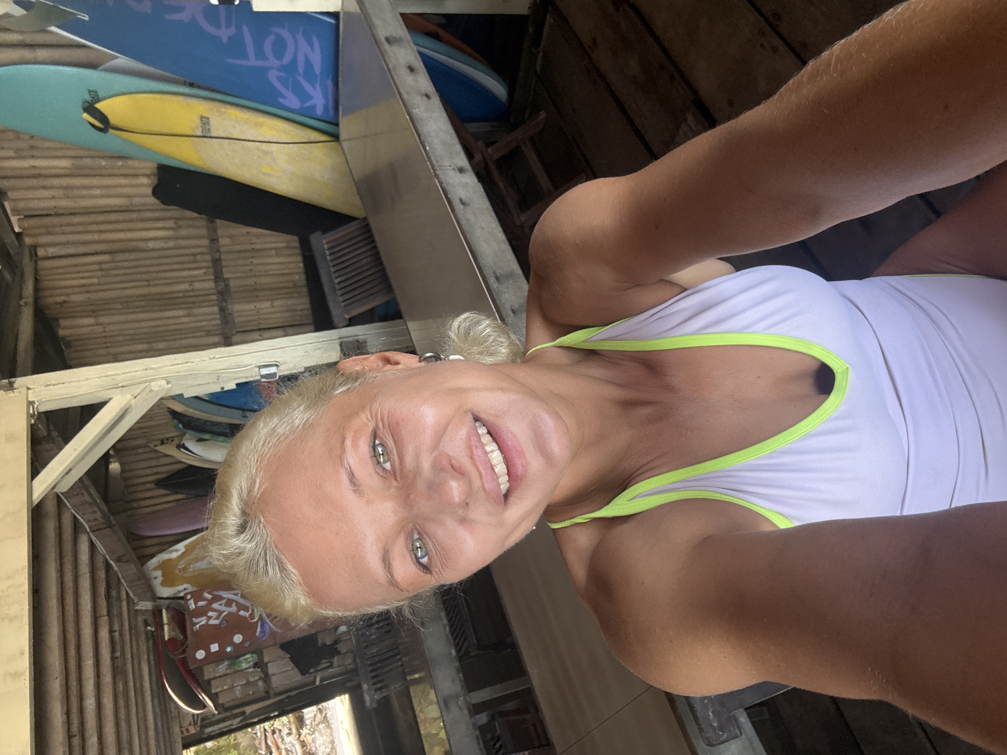 A candid, rotated selfie shows a smiling woman with blonde hair and bright green eyes, wearing a white top with neon green trim. Her face is in the foreground, angled slightly upwards, conveying a cheerful and happy mood.
The background is filled with multiple surfboards stacked or propped up against a rustic wooden structure. Visible surfboards include a prominent blue one with partial text "NOT" clearly visible, a light blue board, a yellow board, and a pink one. The yellow board has a small black graphic or text near its fin. The structure itself consists of wooden planks, beams, and what appears to be part of a wooden slatted chair or bench, suggesting a casual, possibly outdoor or semi-enclosed setting like a surf shack, storage area, or rental shop.
The lighting indicates daytime. The image's orientation is significantly tilted, rotated approximately 90 degrees counter-clockwise.