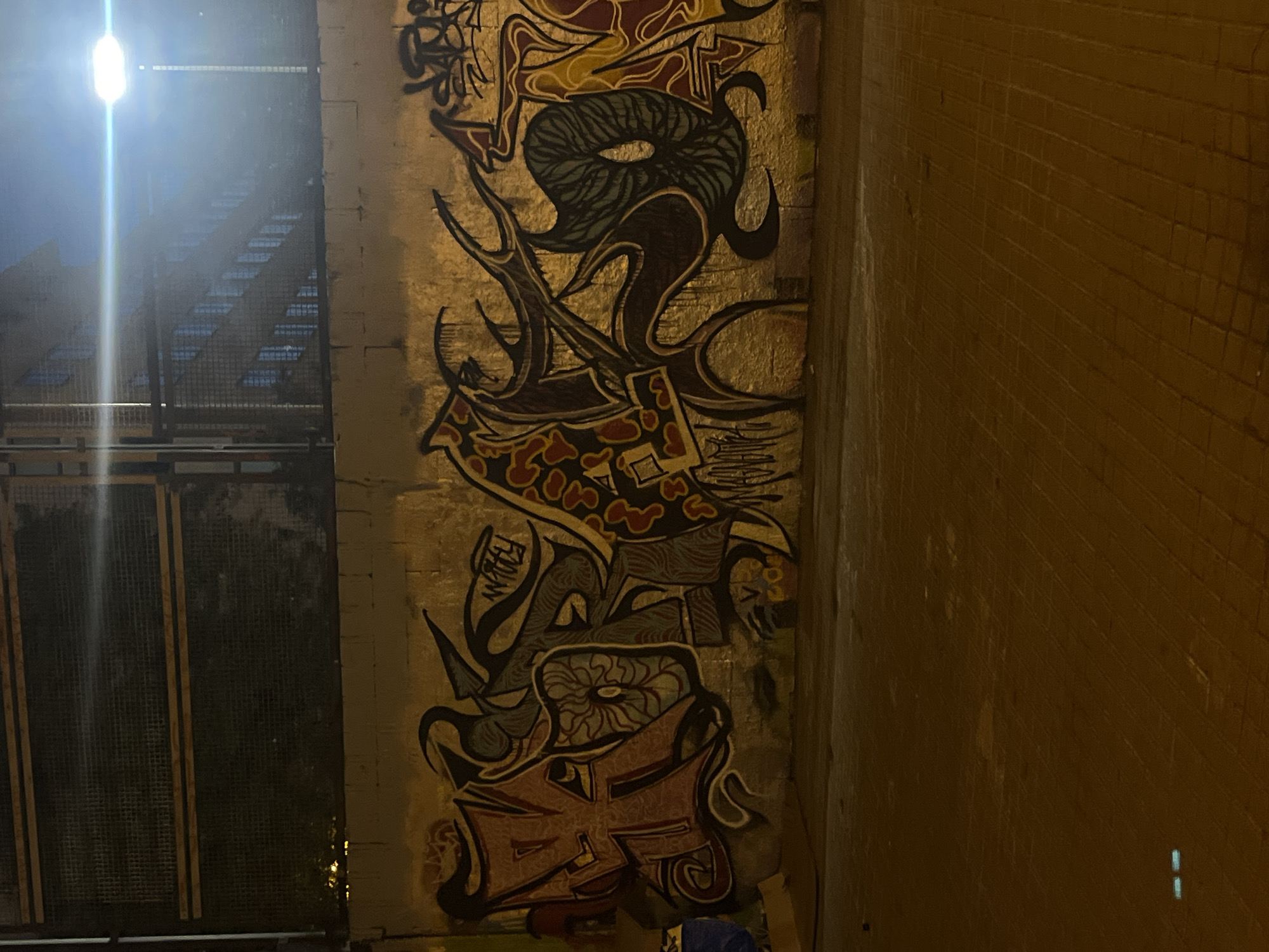 The image depicts a section of an urban environment, likely an alleyway or underpass, at night. A wall covered in graffiti art is the central element, featuring a complex mix of shapes, lines, and colors. Adjacent to the graffiti wall is a dark, metal fence or gate-like structure with a staircase ascending beyond it. The right side of the frame shows a tiled wall. The bright light source above and to the left of the staircase provides the primary illumination. The graffiti itself appears to be a collection of abstract designs. The setting appears to be a street in Barcelona, Spain.